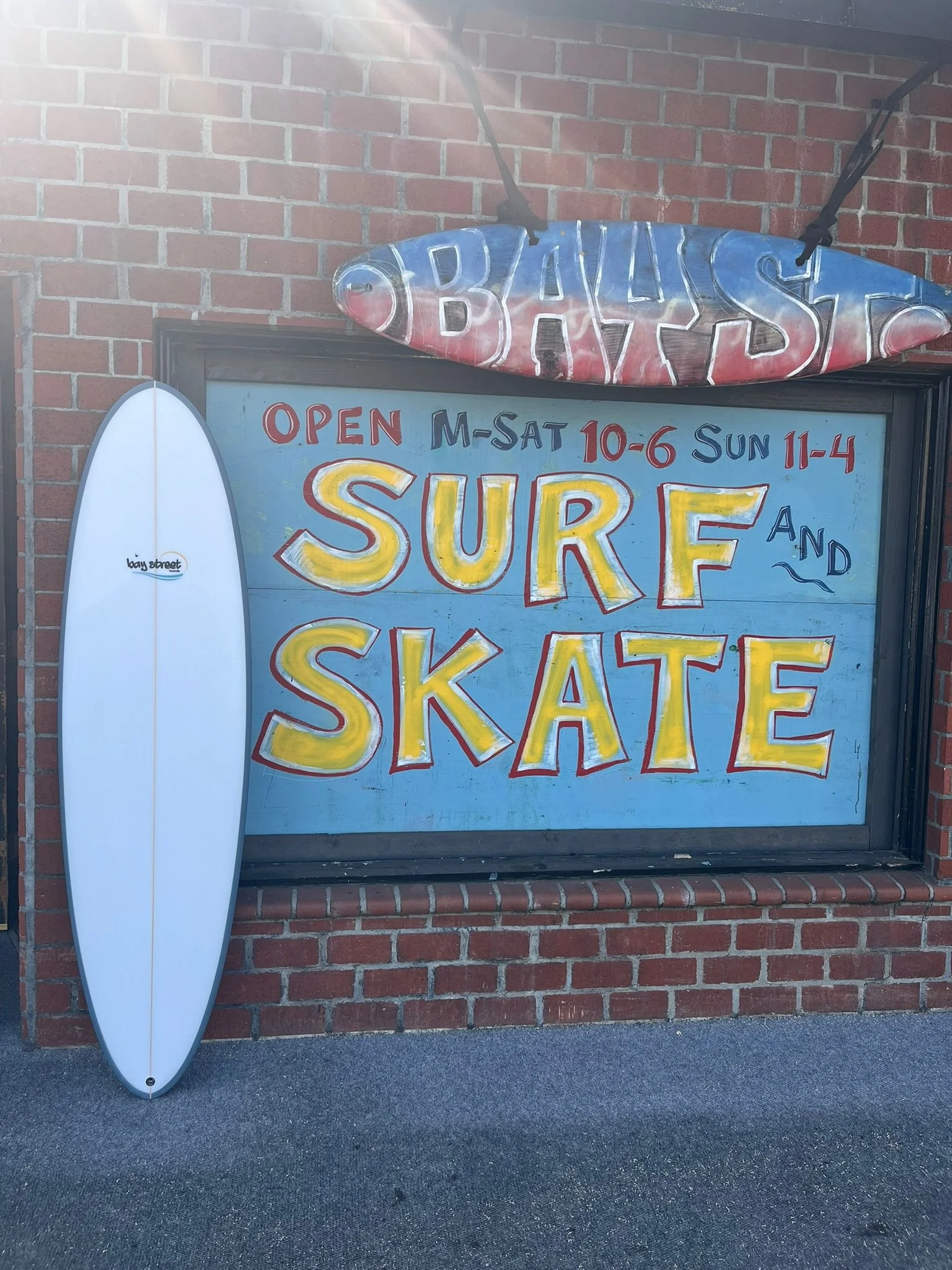 A surfboard leans against a brick wall next to a sign advertising a skate shop, with hours of operation and a large, colorful sign reading 'SURF SKATE SALE' in yellow and red letters.