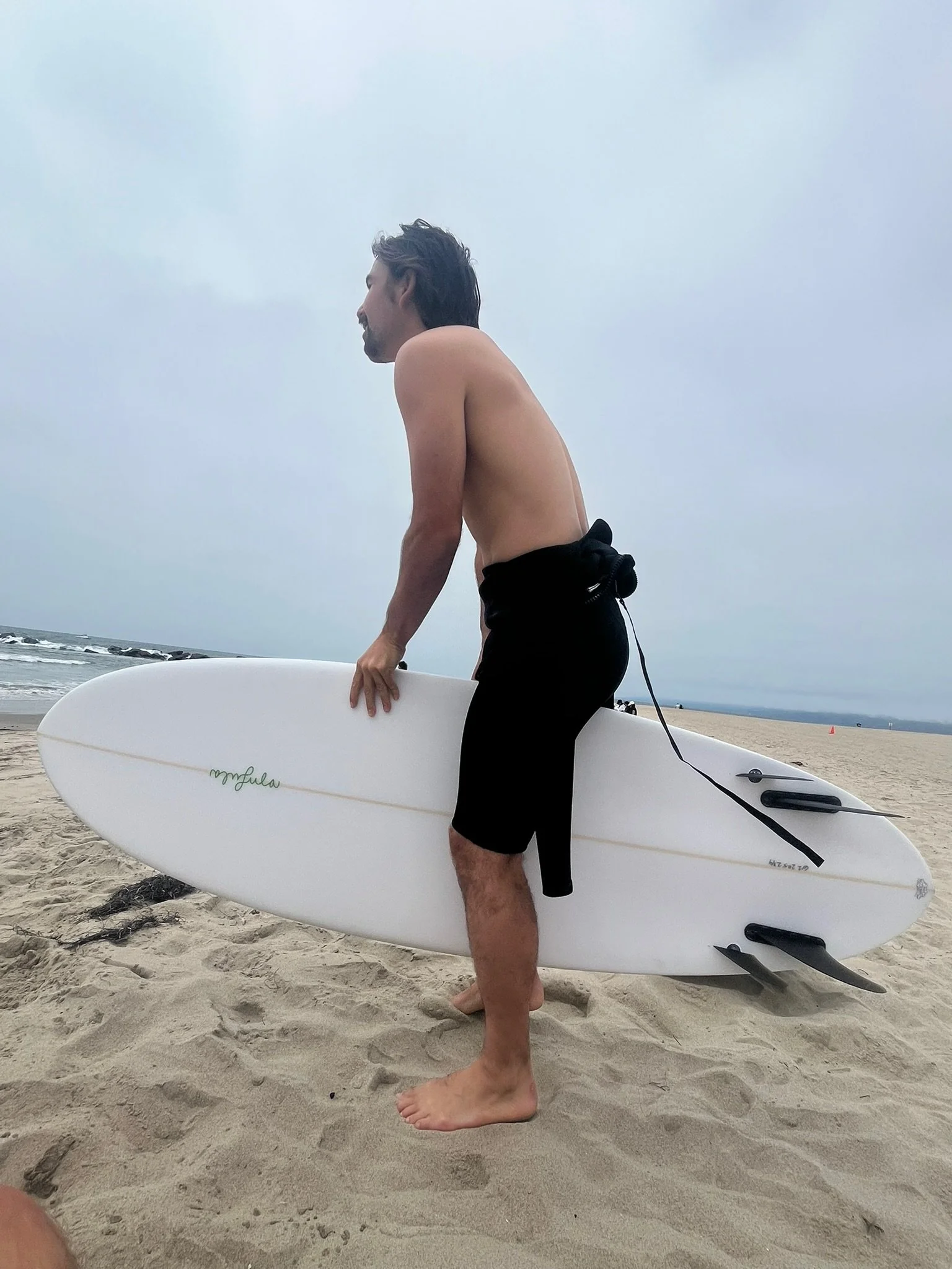 Mollusk surf shop employee with dark hair and a mustache, wearing black shorts, stands on a sandy beach holding a white surfboard, with the ocean and cloudy sky in the background.