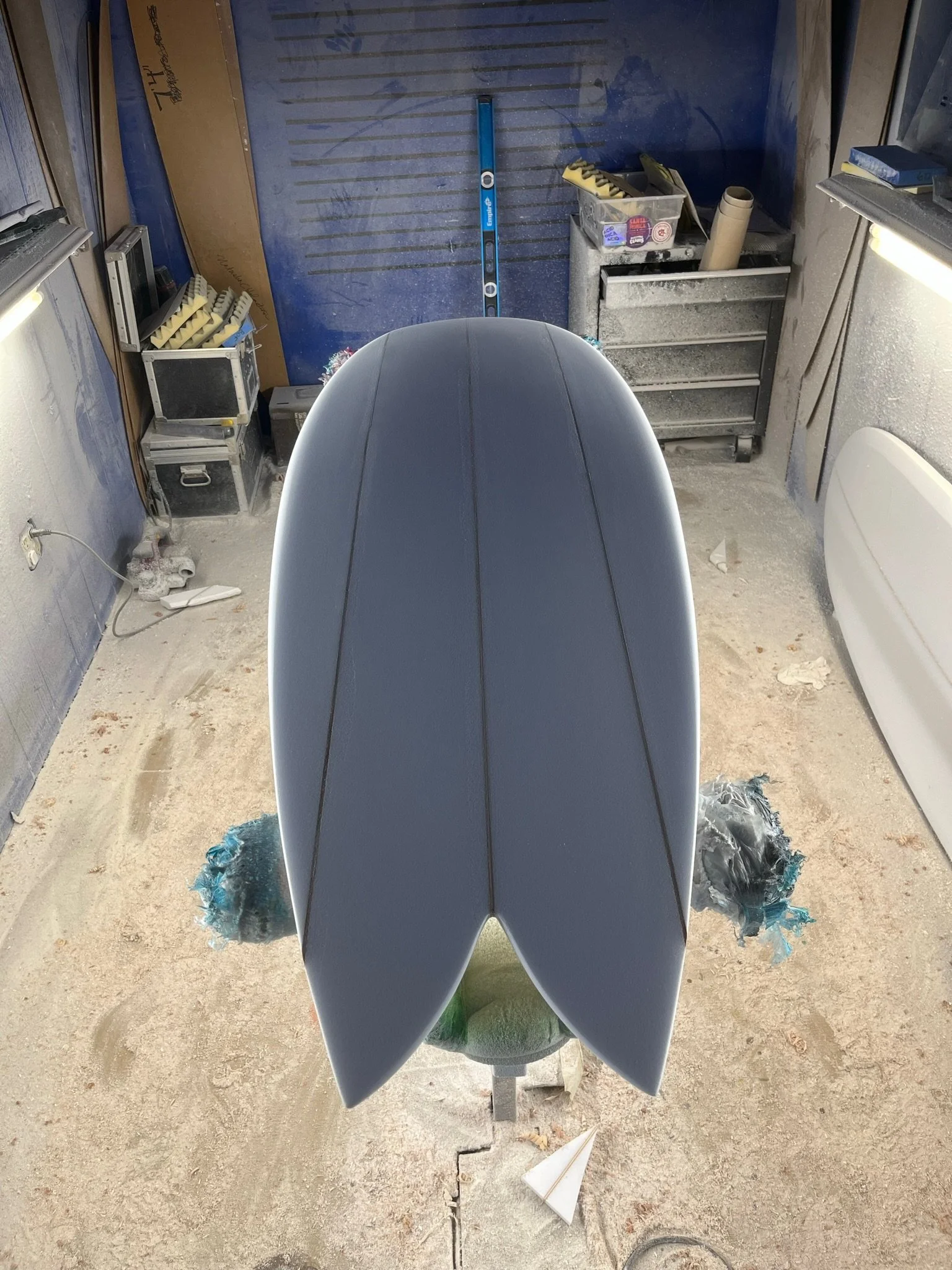 A surfboard with a blue and black design mounted on a stand in a cluttered workshop space, with tools, boxes, and construction materials surrounding it.