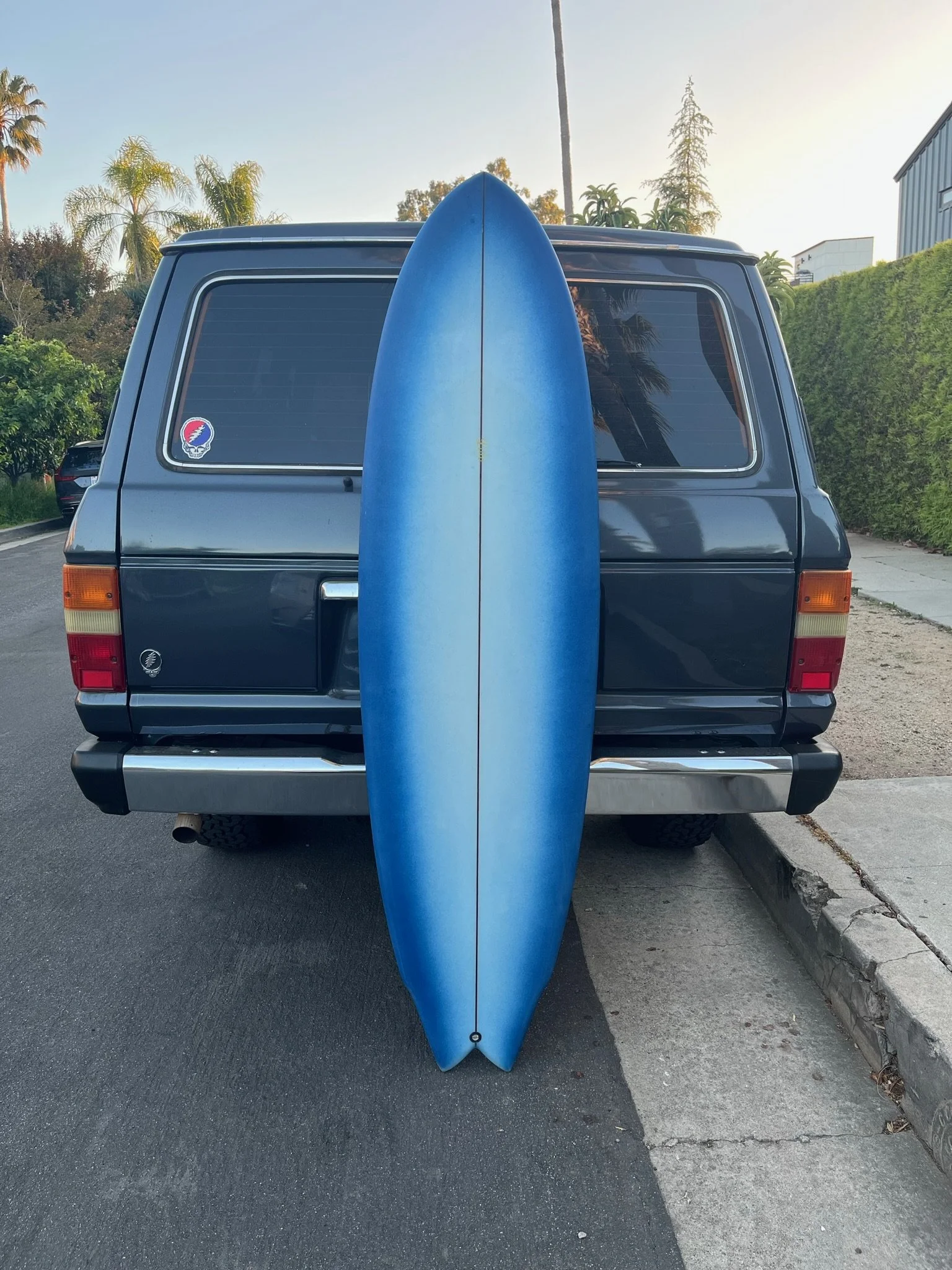 Blue surfboard standing upright against the back of a dark gray van parked on the street, with trees and a building in the background.