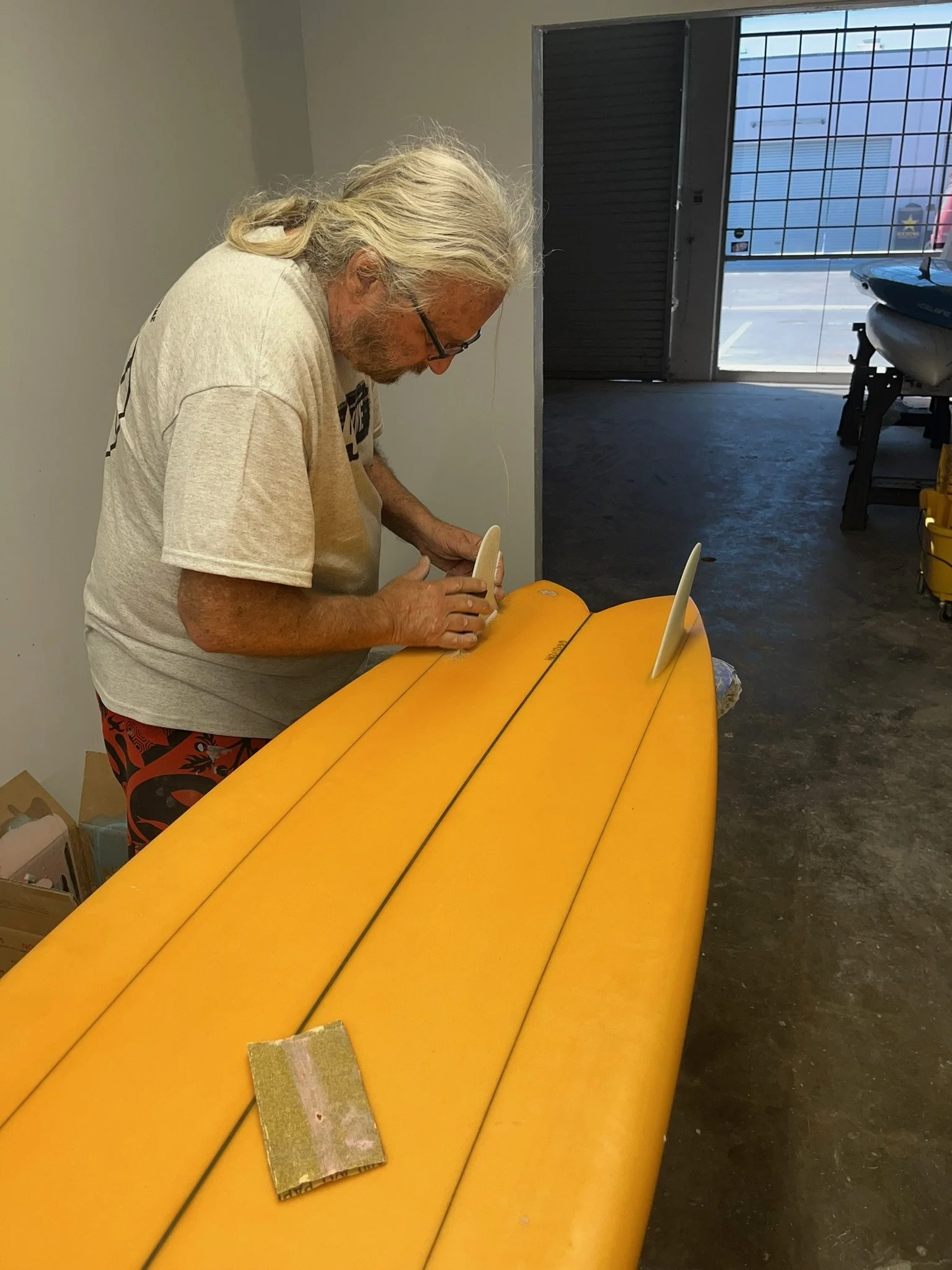 An elderly man Larry Allison fellow surfboard designer, with long gray hair and glasses working on a yellow surfboard indoors, with a garage door in the background.