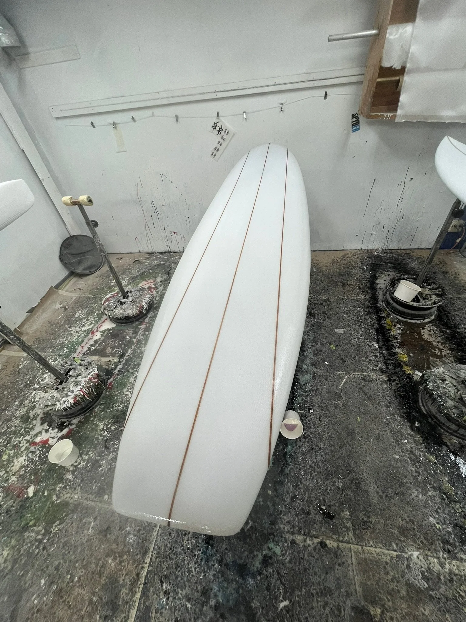 A surfboard in a cluttered, dirty workshop or garage with paint splatters, cups, and tool stands around.