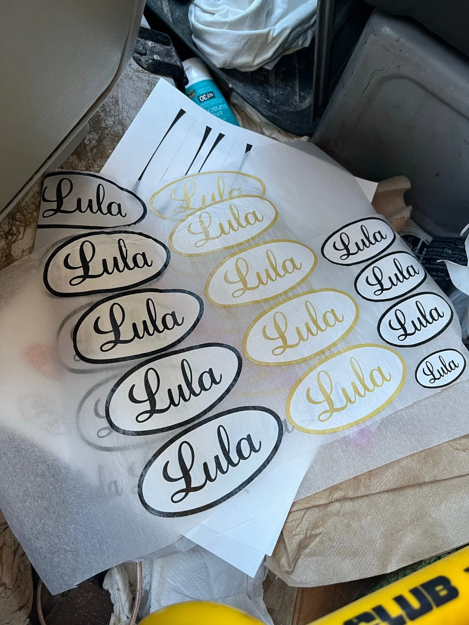 Collection of stickers with the word 'Lula' in black and gold, placed on different sheets of paper near various objects including a container of disinfectant spray and a plastic bag.