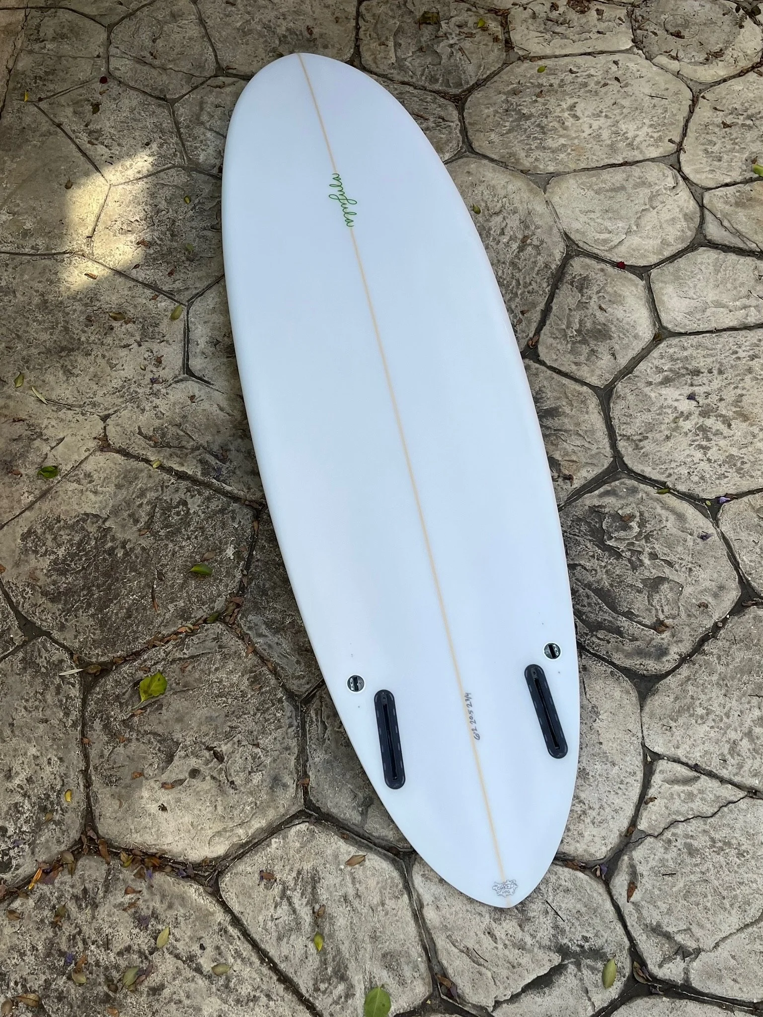 A white quad surfboard rounded pin tail, lying on a stone pathway, with black foot straps and a small logo near the bottom.
