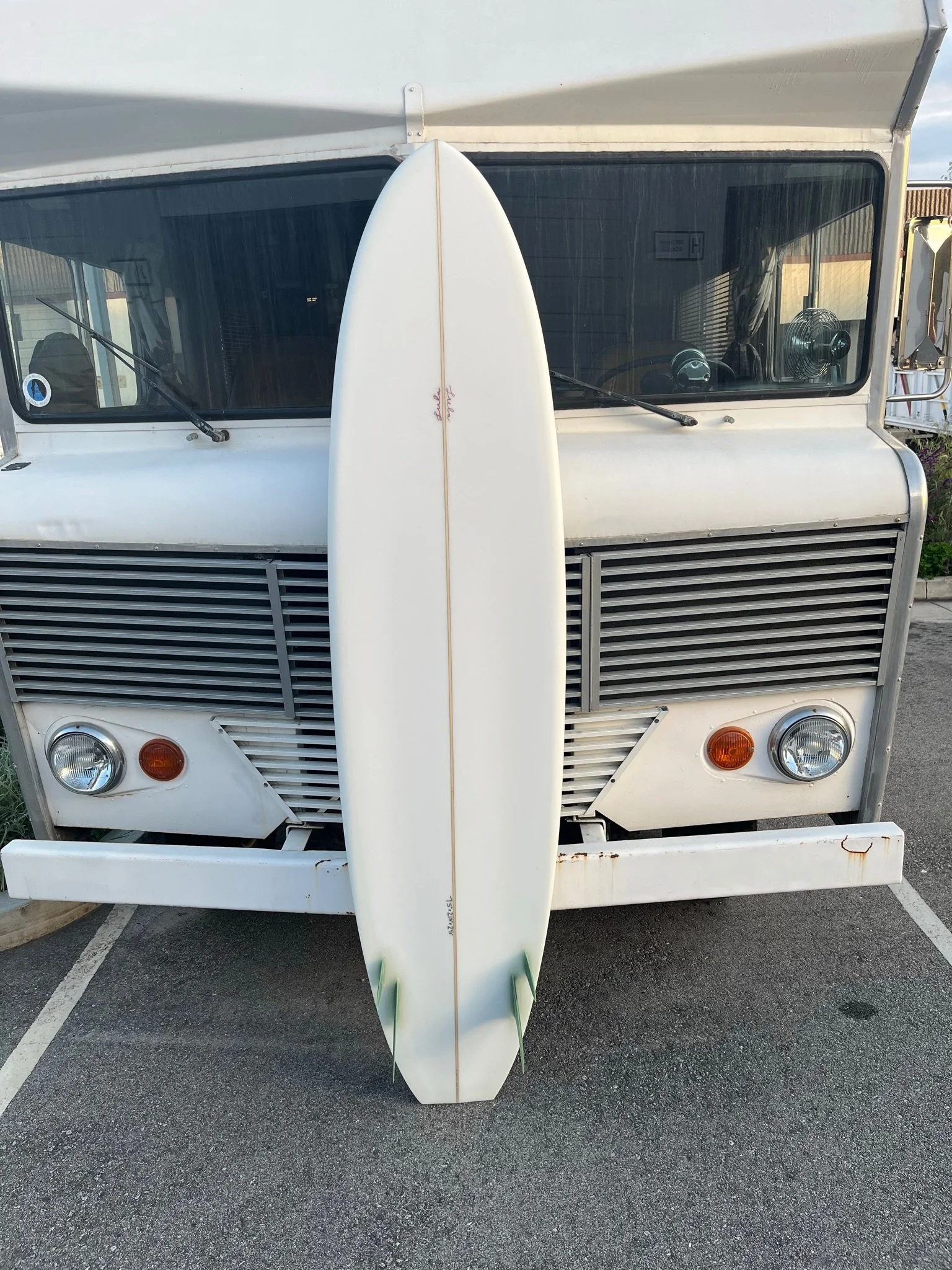 A white surfboard standing upright in front of the front of a vintage camper van, parked in a parking lot.