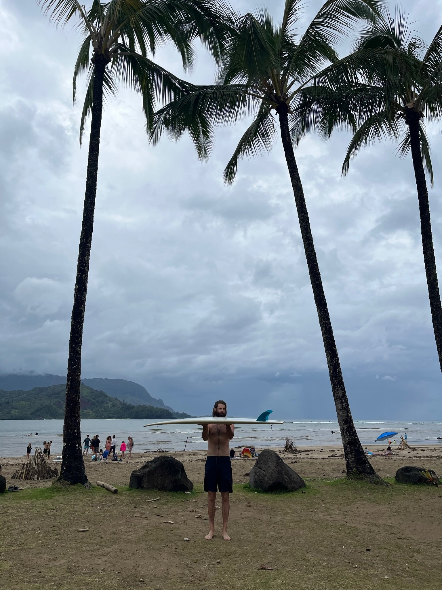 A man holding a surfboard stands on a beach with palm trees and a cloudy sky. People are visible in the background by the water, some with umbrellas and others walking along the shore.