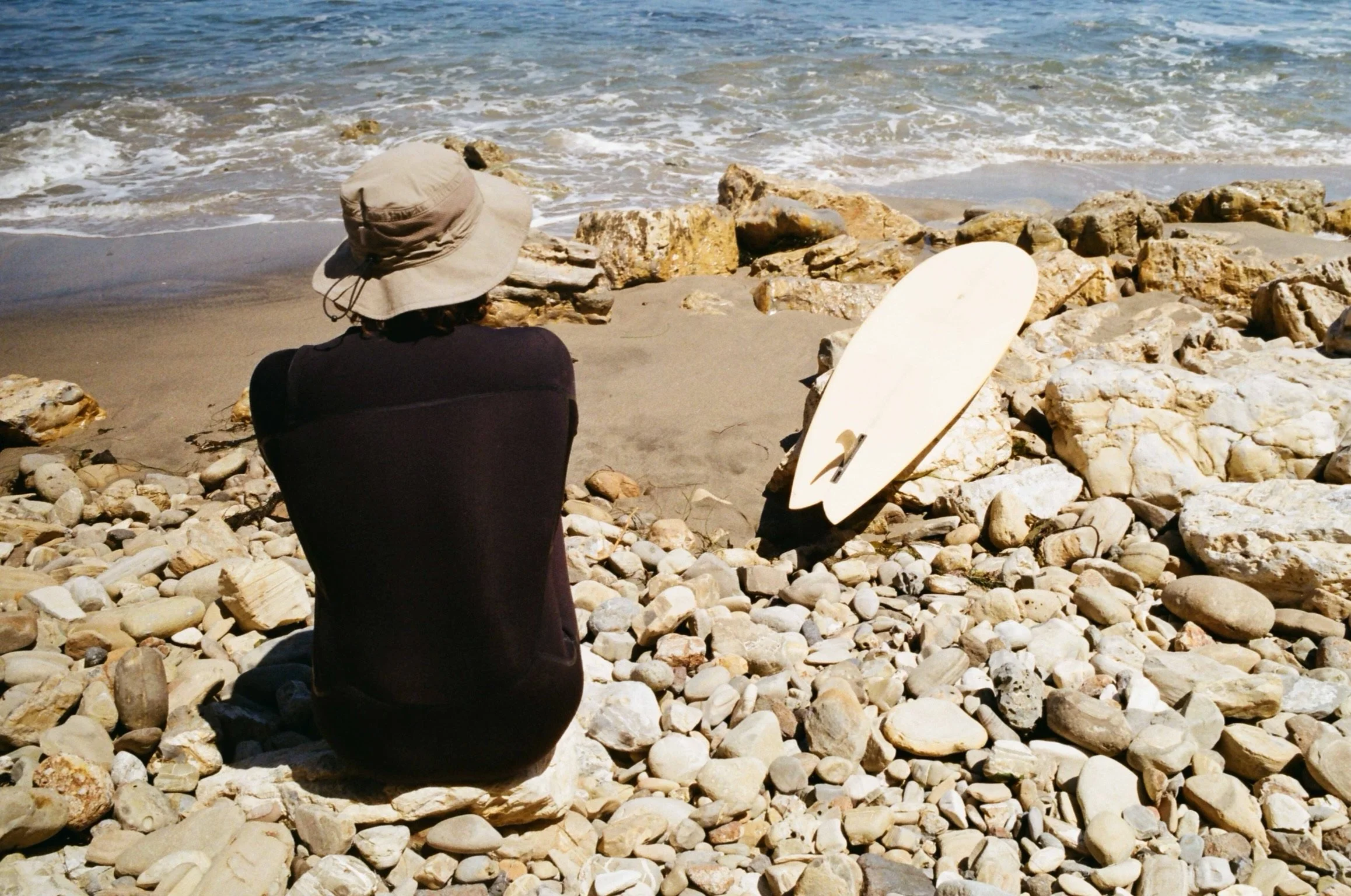 Person sitting on pebble beach facing the ocean with a surfboard.