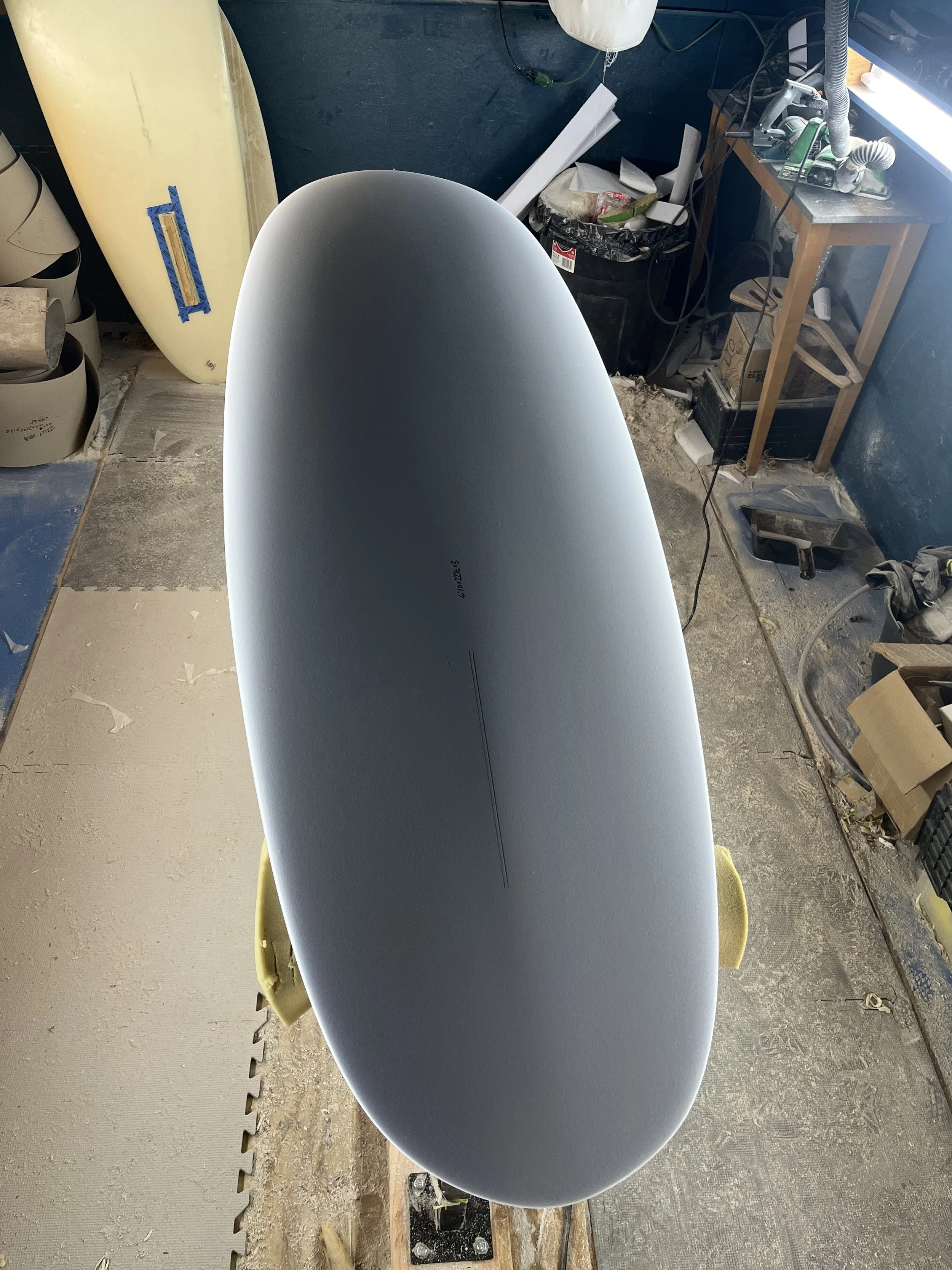 A surfboard resting on a yellow foam block in a workshop with a workbench, tools, and clutter, including a trash can, boxes, and cords.
