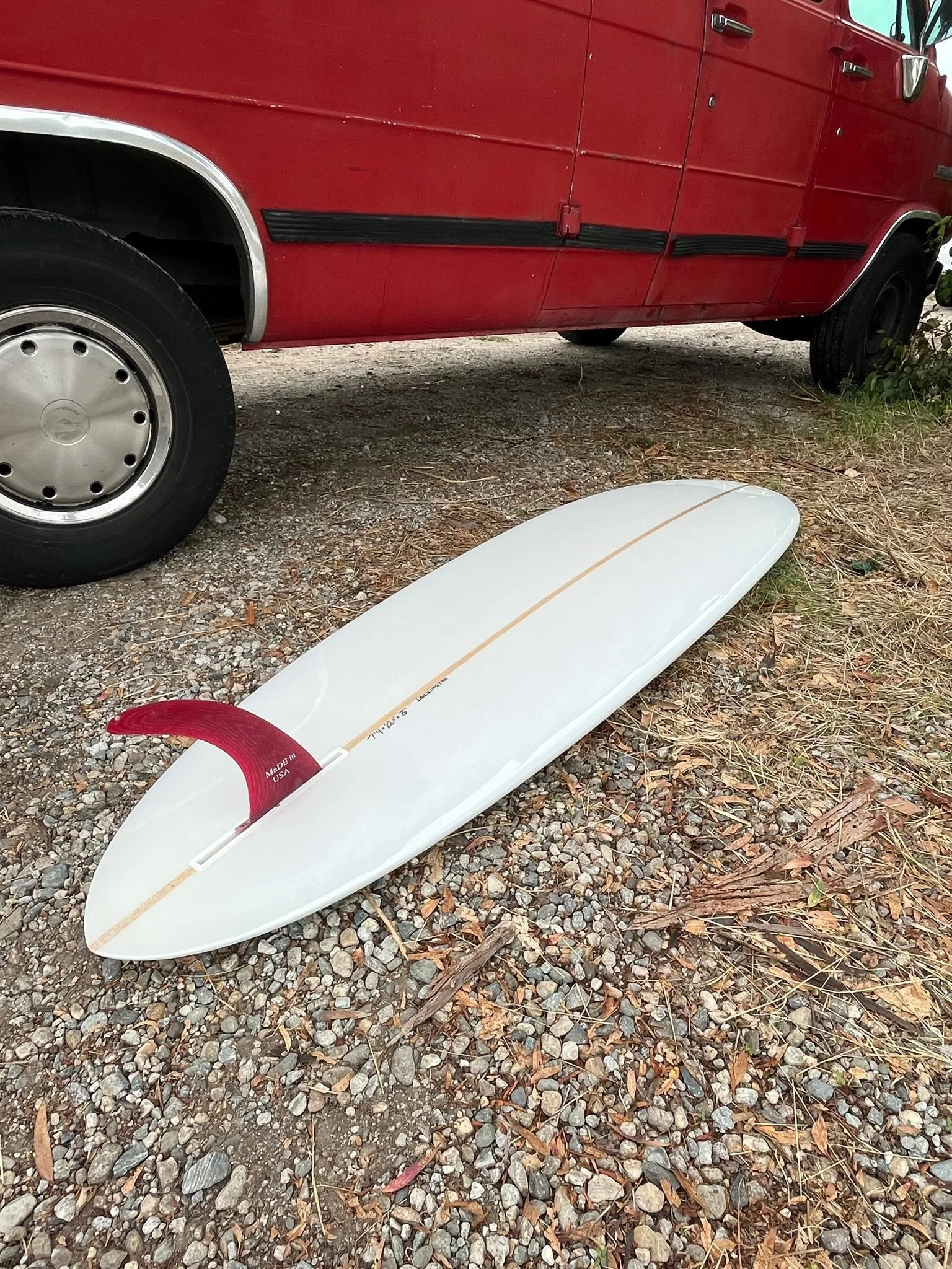 A white surfboard with a red fin lying on gravel ground next to a red vehicle, possibly a van or bus, with black trim and tires.