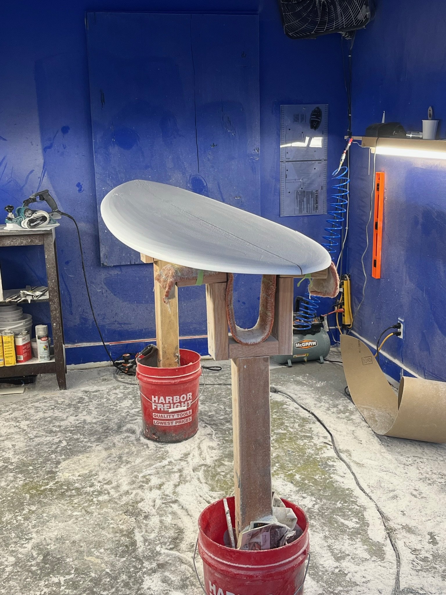 Interior of a workshop with a surfboard on a stand, surrounded by tools, equipment, and a blue wall, with a red bucket on the floor