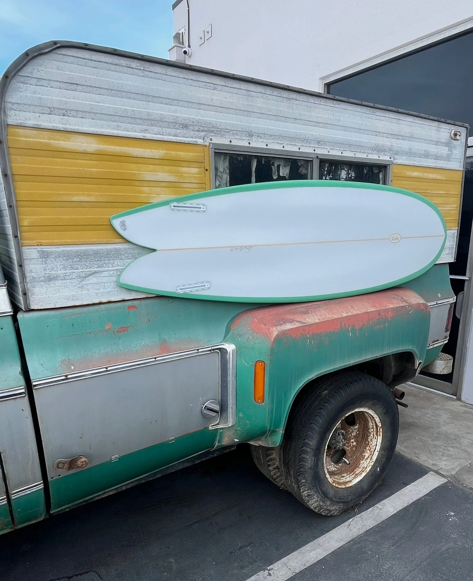 An old green and yellow camper van with a surfboard on the side, parked in a parking lot. The van has rusty wheels and a weathered appearance.