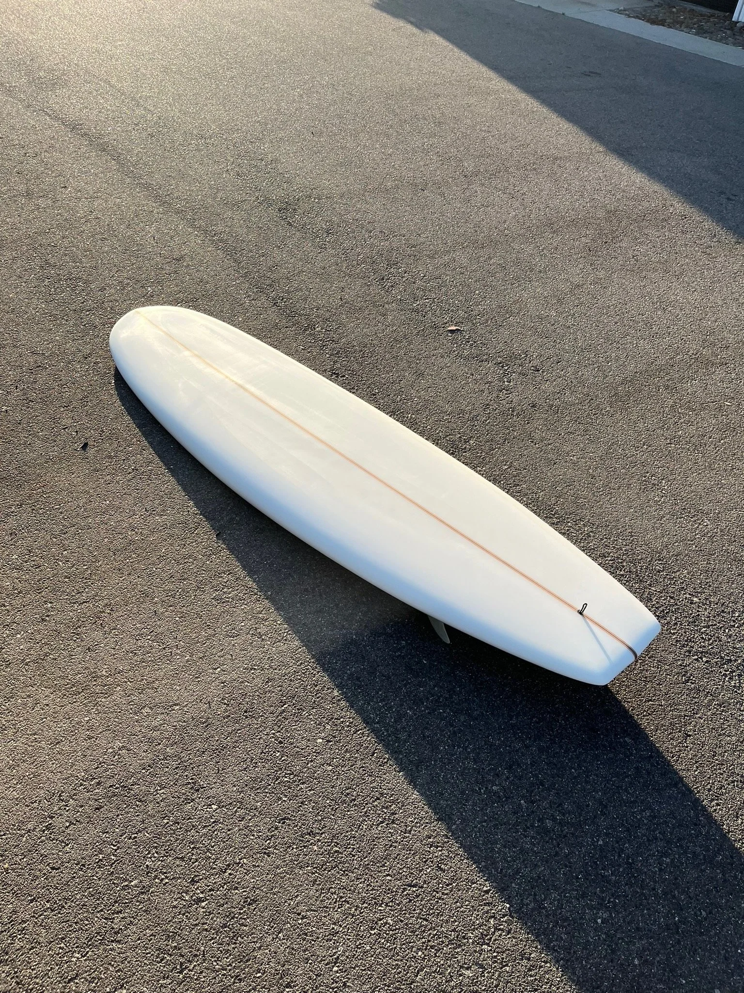 A white surfboard lying on dark asphalt near a curb with a shadow cast nearby.