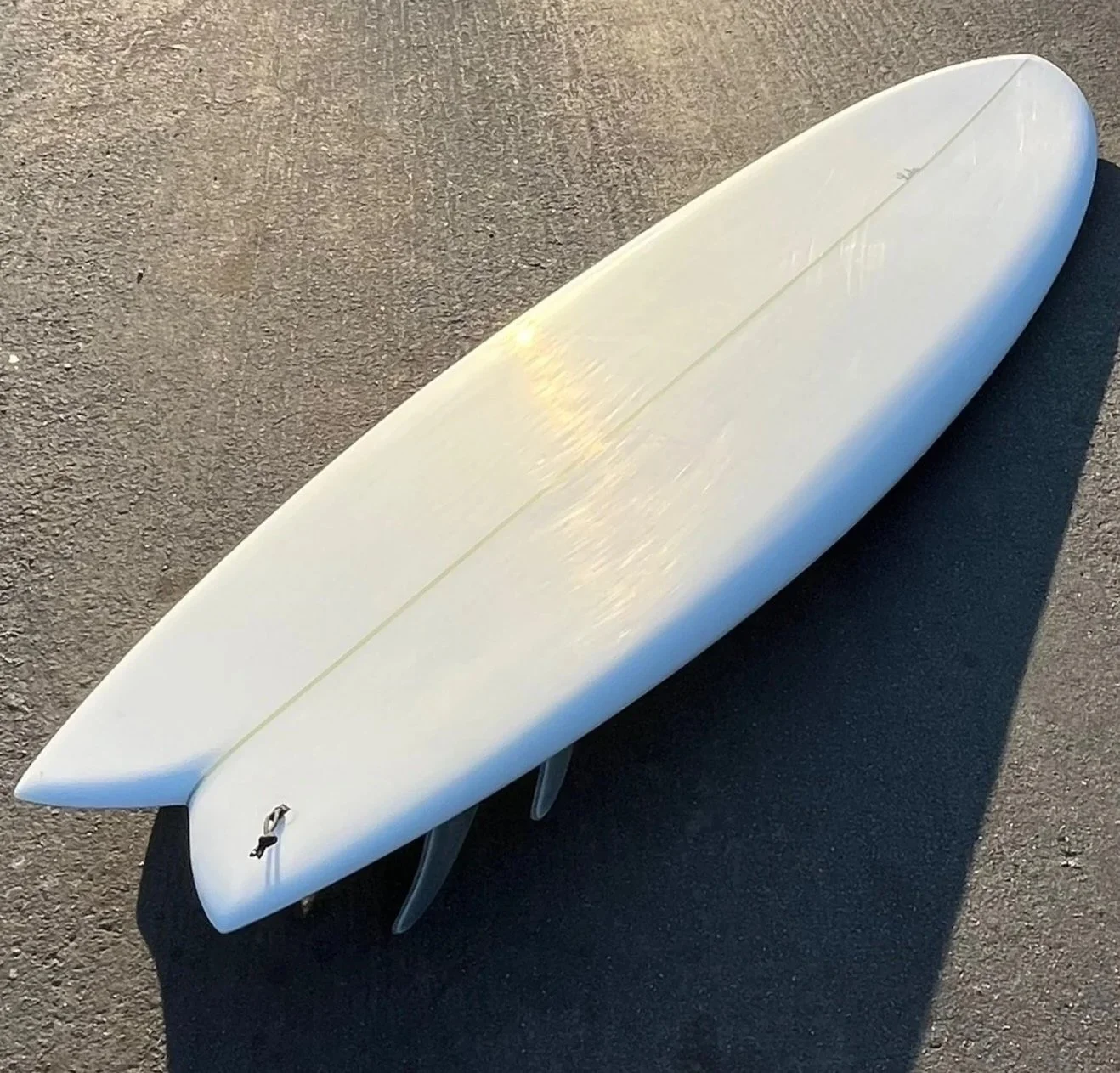 White surfboard with three fins on a paved surface.