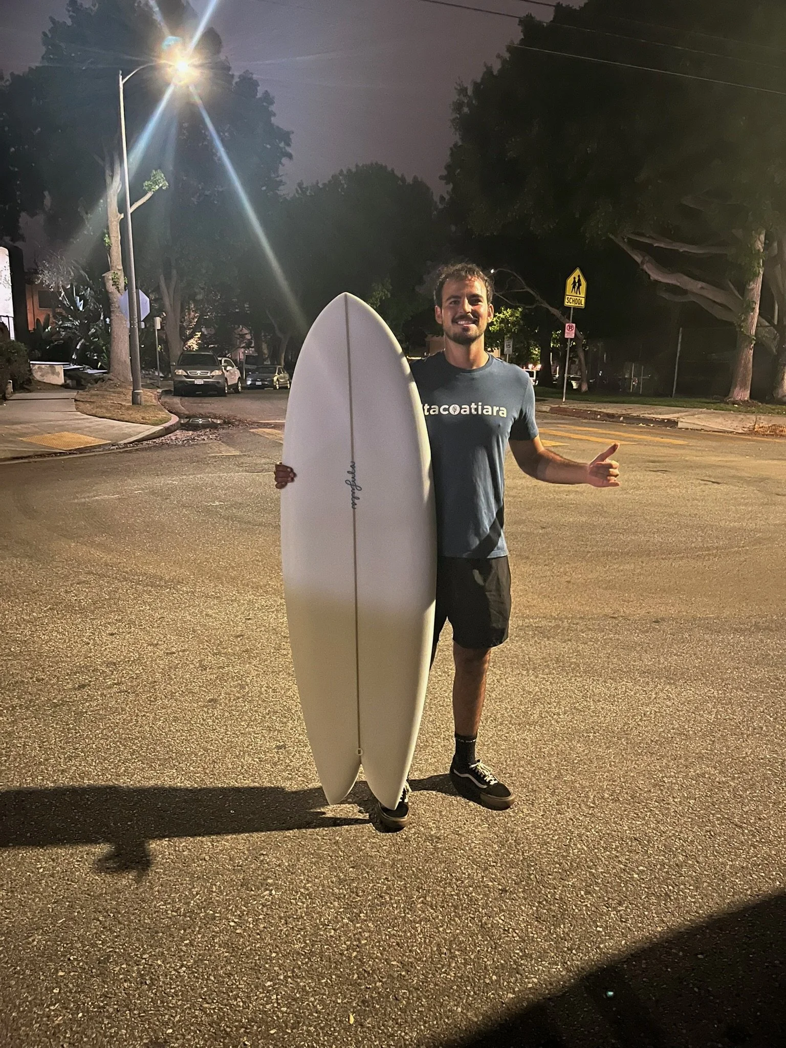 A man standing on a street at night holding a surfboard and giving a thumbs-up. He is dressed in a t-shirt, shorts, and sneakers, smiling. The background shows parked cars, a school zone sign, trees, and streetlights.