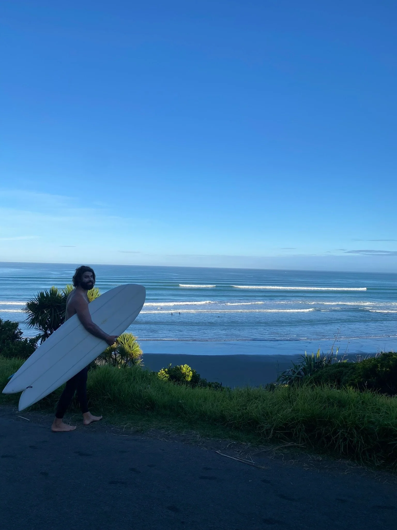 A man holding a surfboard standing on a grassy area near the beach with the ocean in the background and blue sky above.