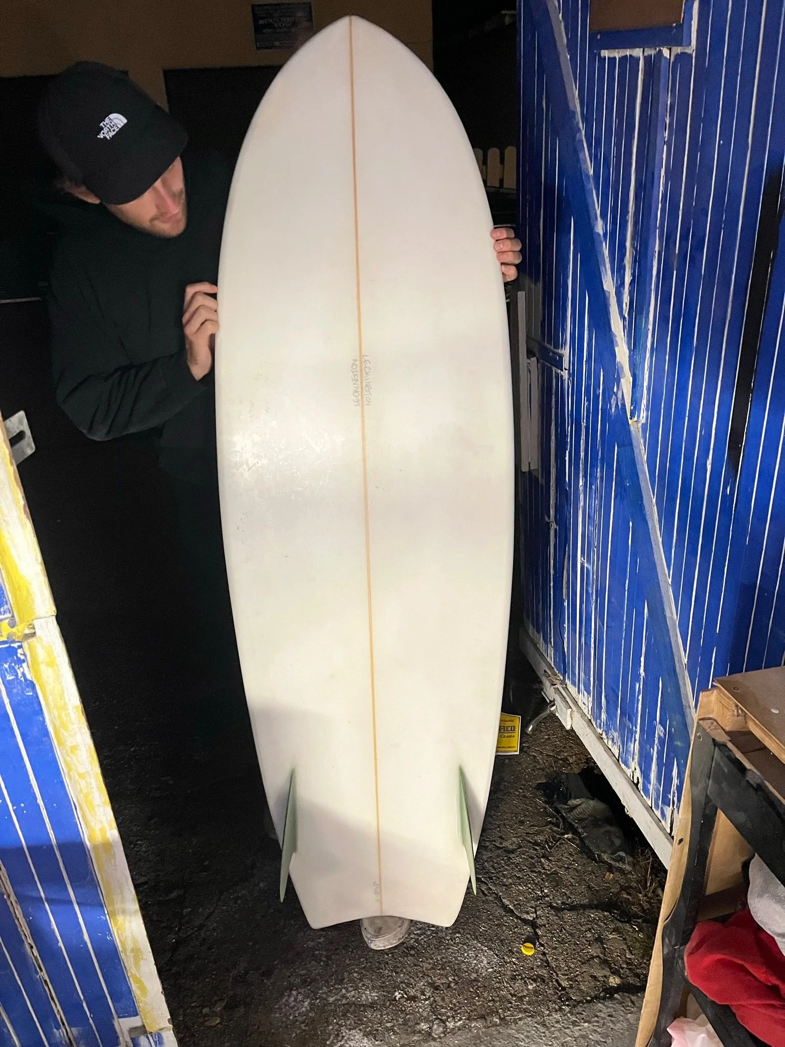 A person in a black jacket and black cap kneeling behind a white surfboard with a yellow strip and text on it, in a dimly lit room with blue and yellow lockers.