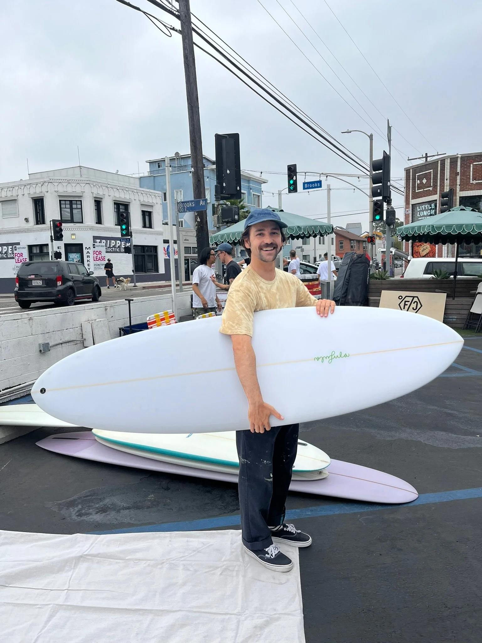 A man with a mustache, wearing a blue cap and yellow tie-dye shirt, standing in a parking lot holding a white surfboard. There are additional surfboards stacked on the ground behind him, and people walking in the background with storefronts and traff
