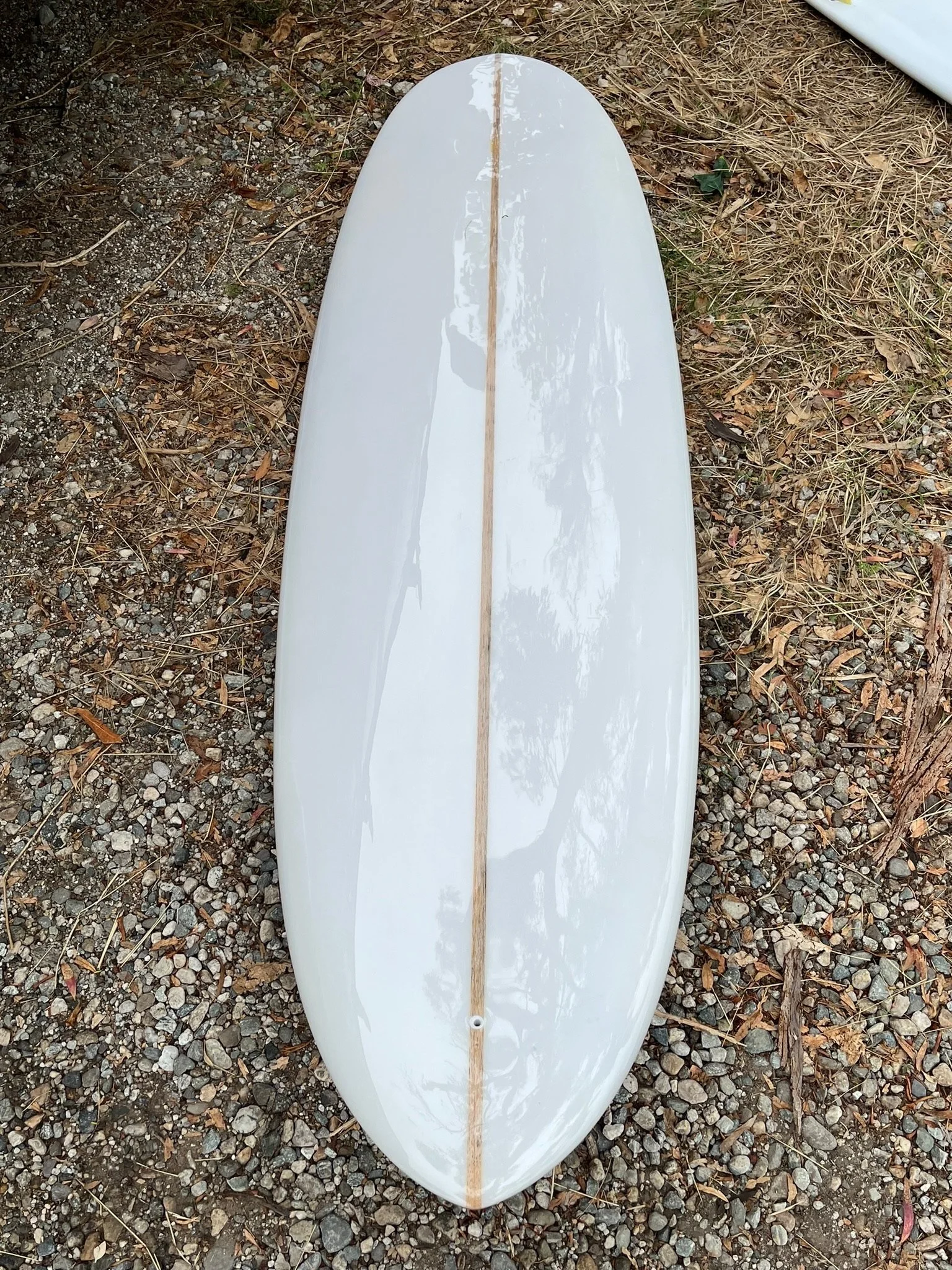 A white glossy surfboard with a brown stringer down the middle, lying on gravel and dirt ground.