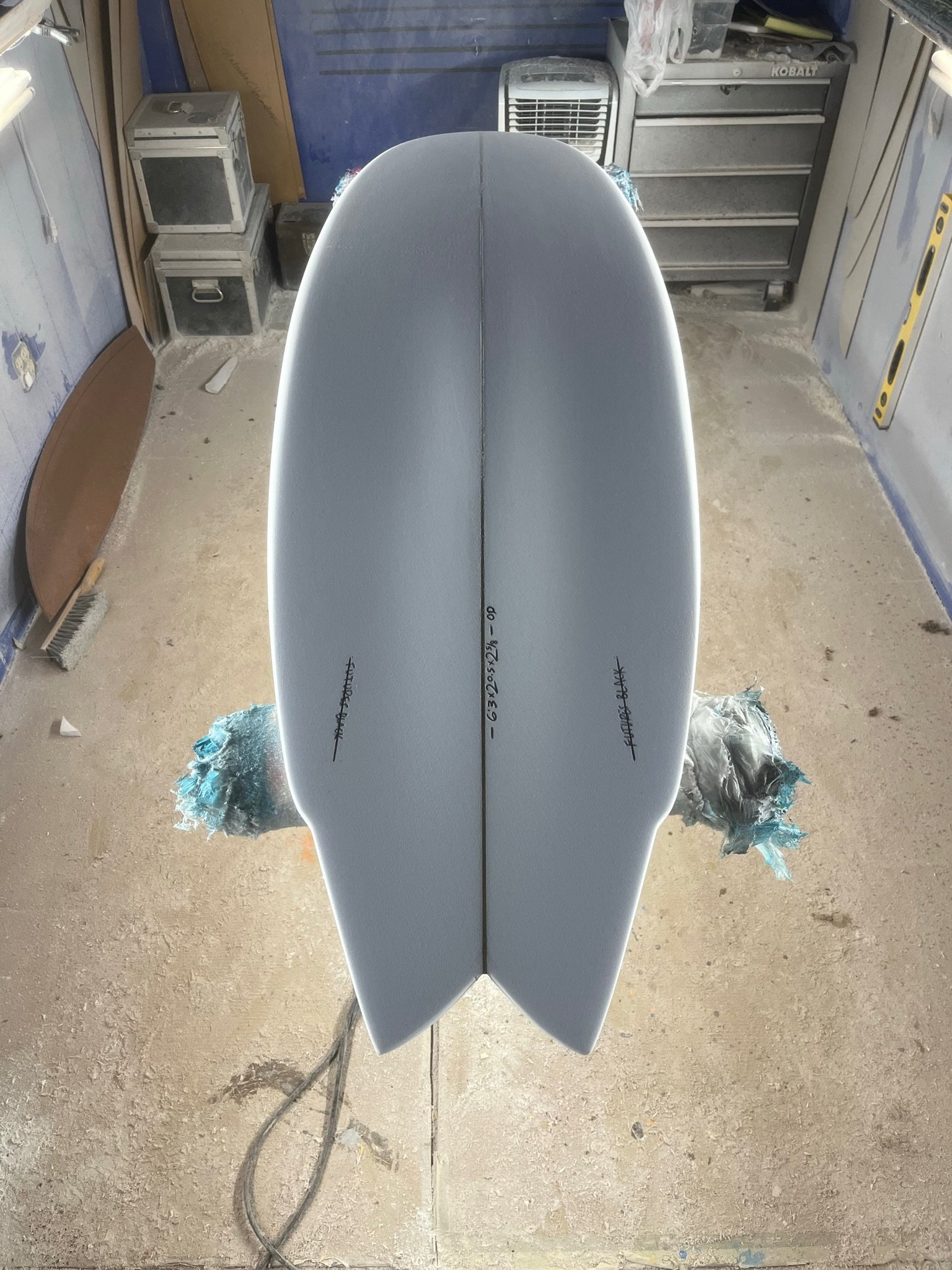 A surfboard with a white top surface and black handwritten measurements written on it, leaning against a wall in a workshop or garage. The background has various tools, storage drawers, and a small electric fan. The floor is dusty.