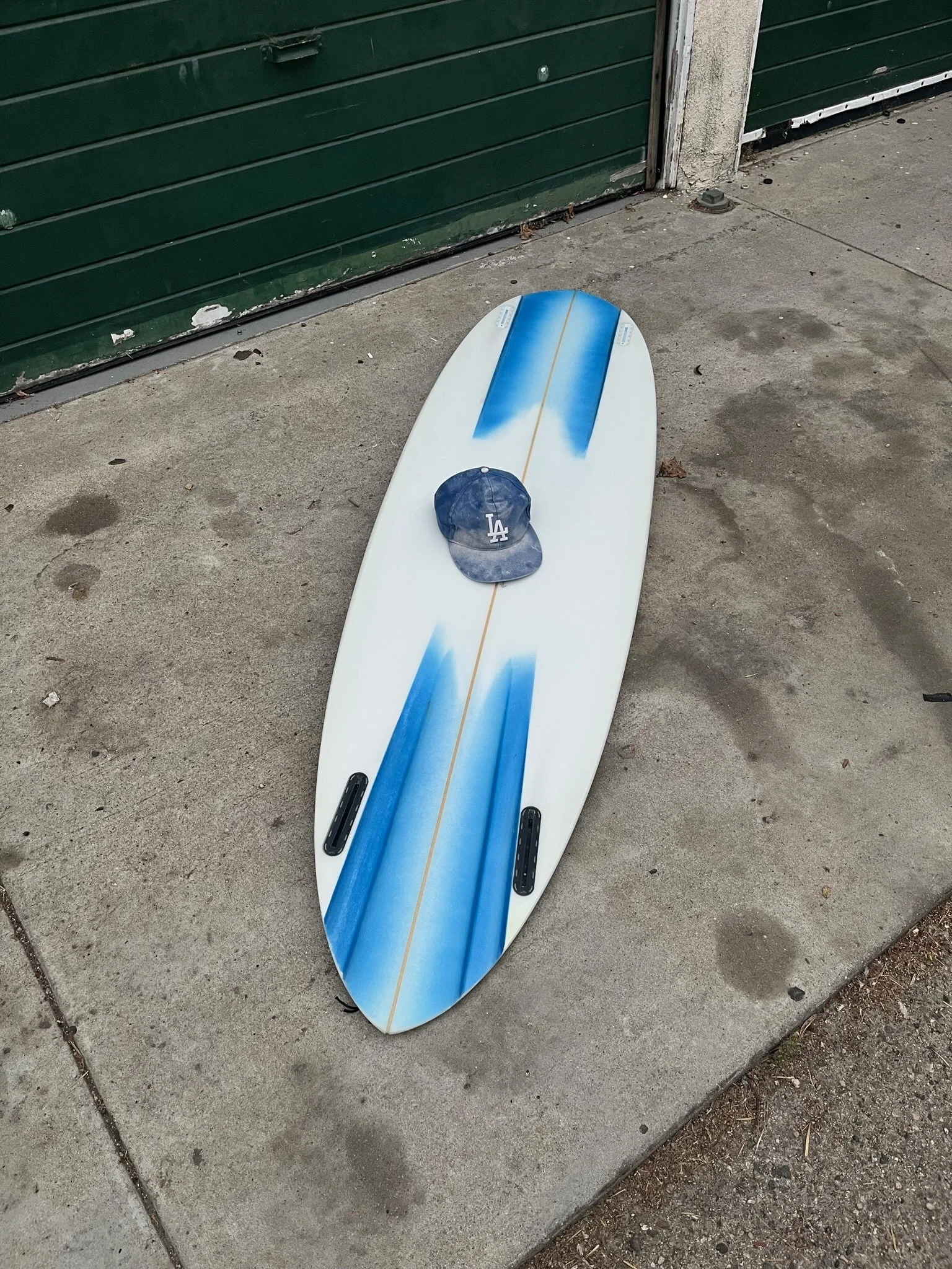 A unique designed surfboard with a white and blue design lying on the sidewalk next to a green metal door, with a dark Los Angeles Dodgers baseball cap placed on it in Venice California.