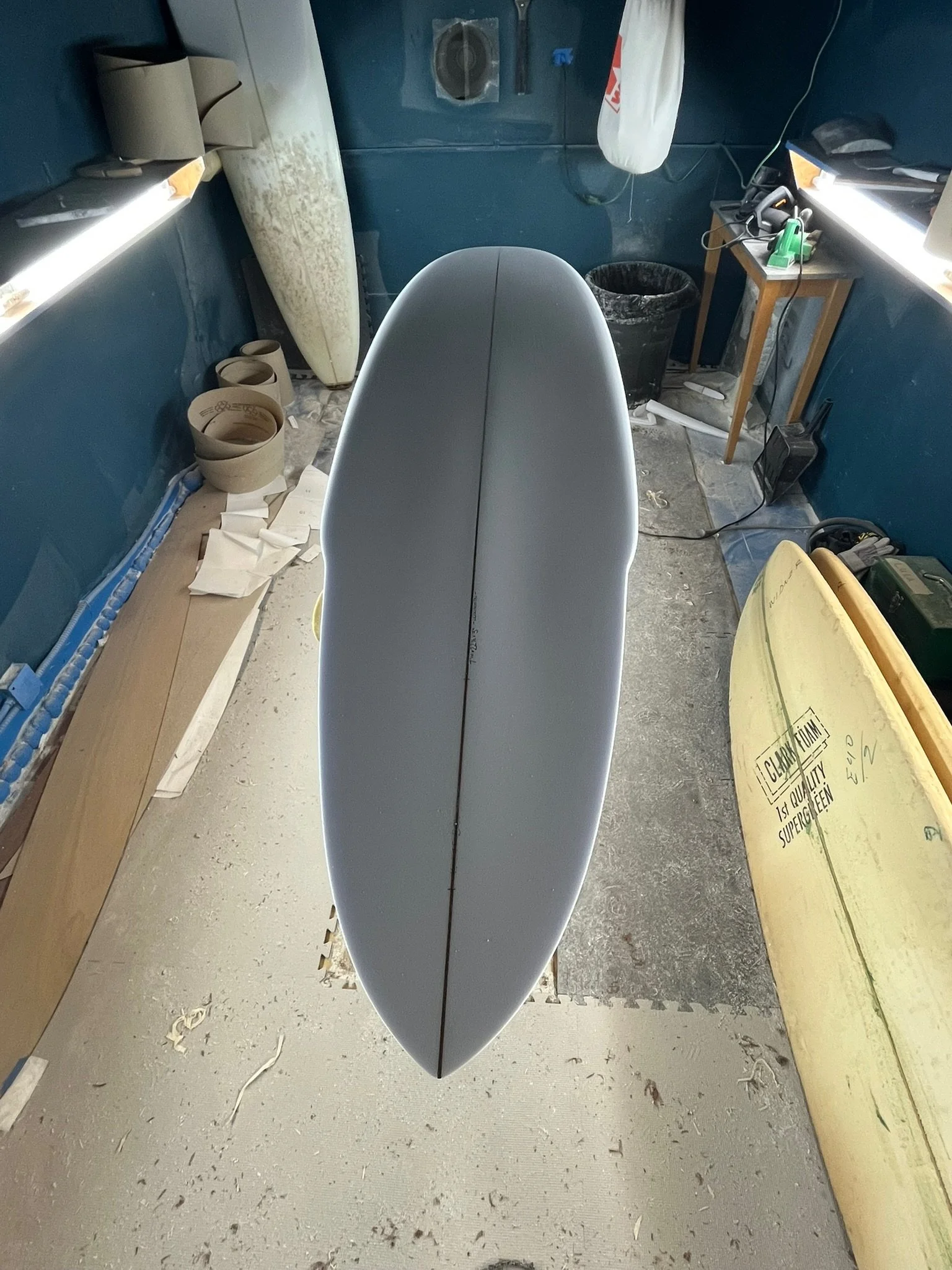 A photo of a surfboard inside a workshop, viewed from above, with various equipment and materials around, including a trash bin, a small table with tools, and a partially visible yellow surfboard on the right.