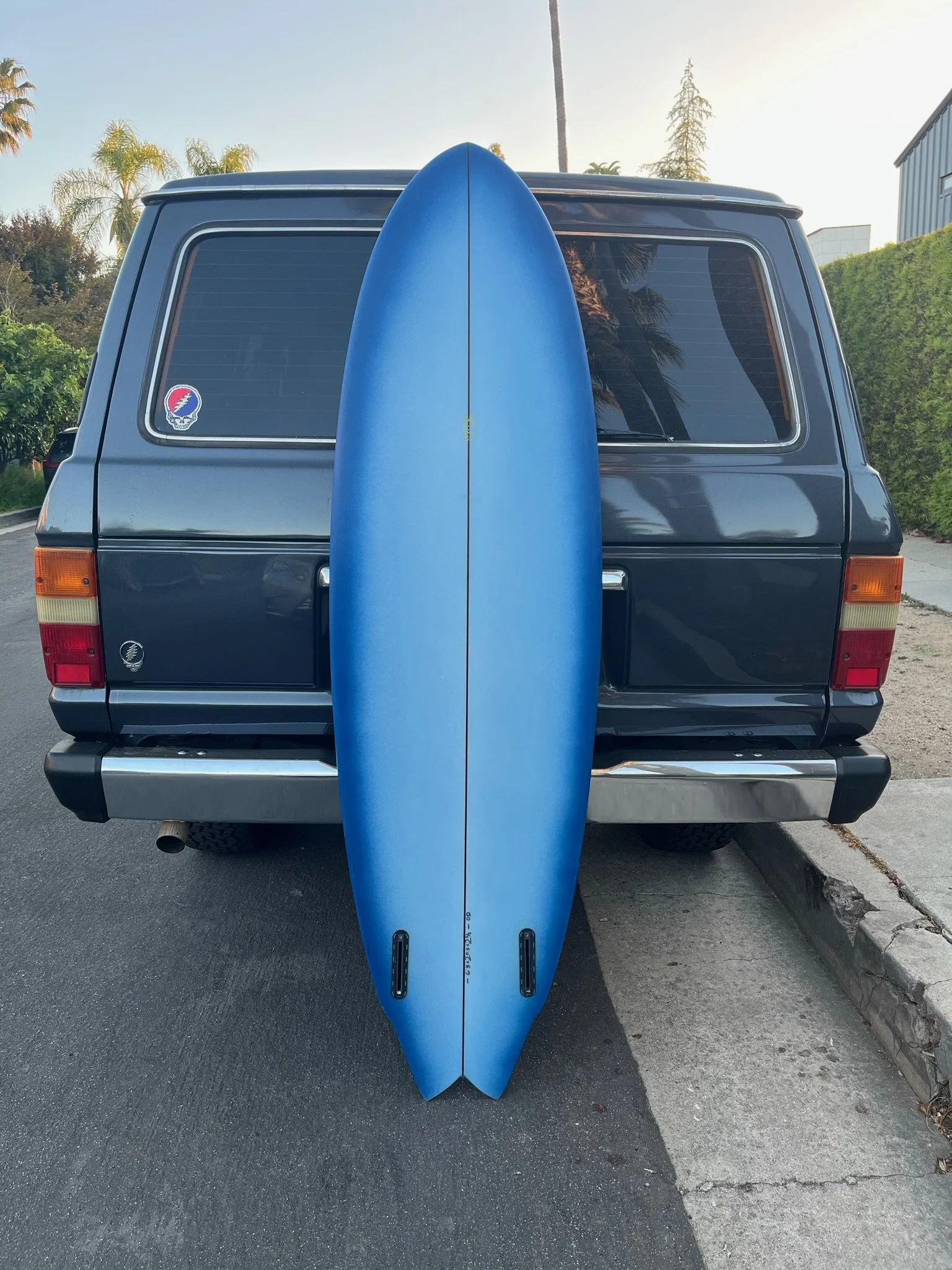 Blue surfboard standing upright behind the back of a dark gray van parked on the street.