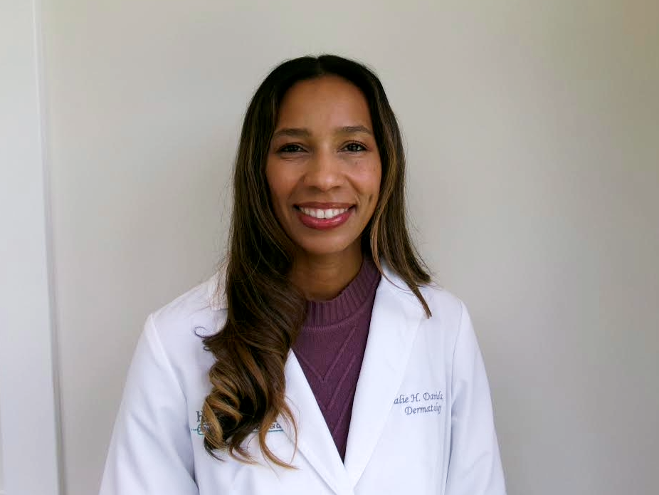 A smiling woman with long brown hair in loose waves, wearing a white medical coat and a purple sweater, standing against a plain light-colored wall.