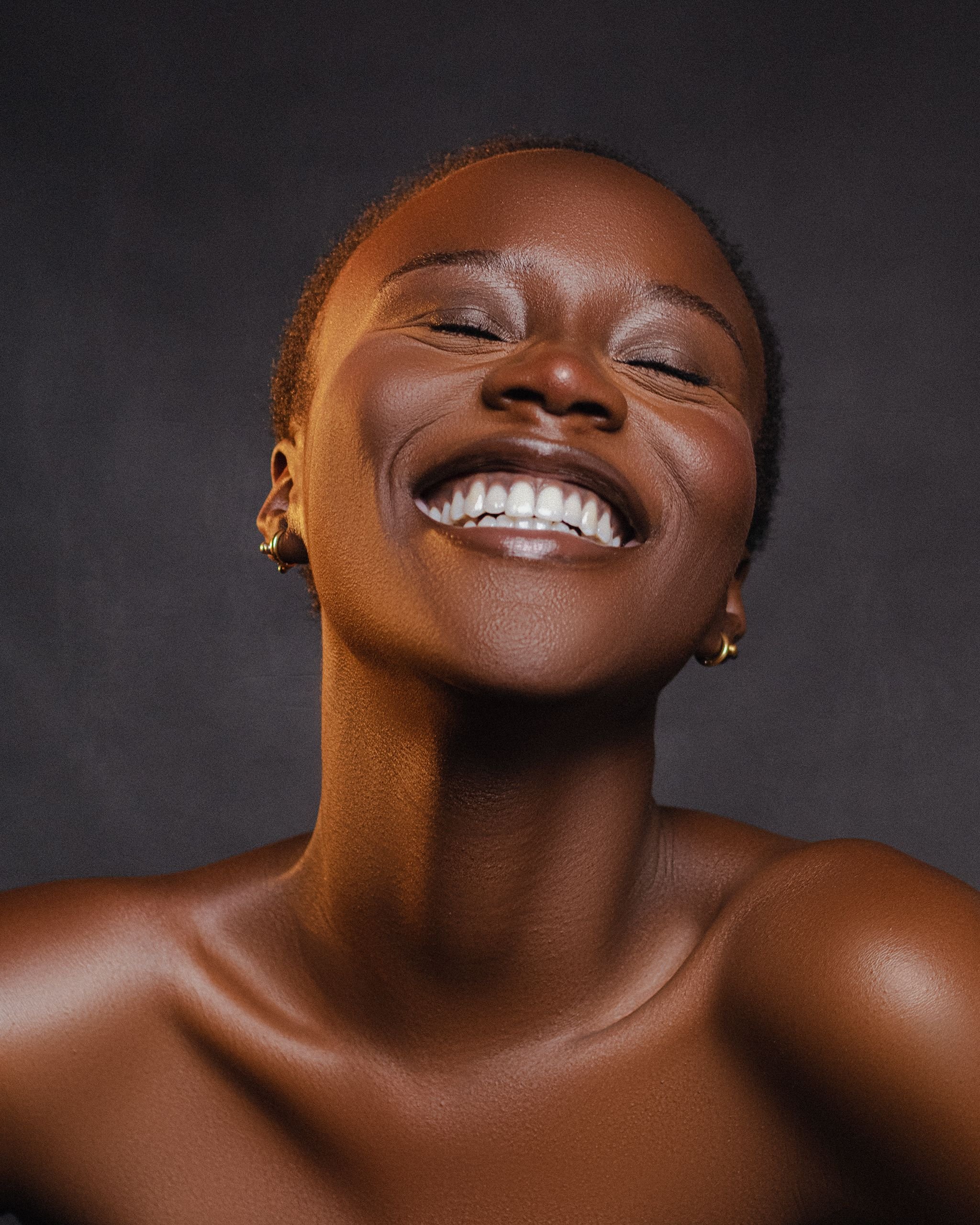 Close-up of a smiling woman with short curly hair, gold earrings, and smooth brown skin, against a dark background.