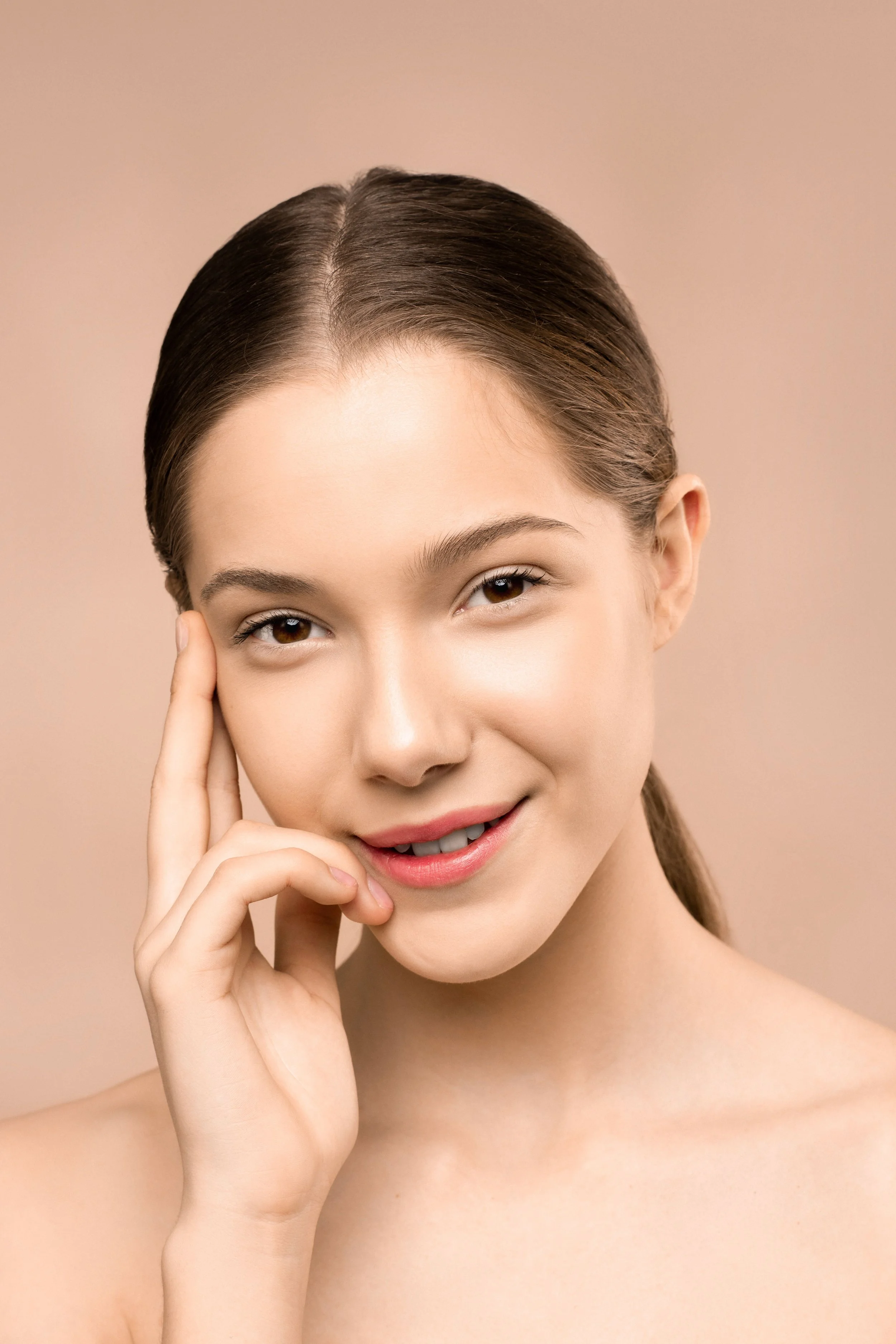 A young woman with brown hair styled back, smiling softly, touching her face with her left hand, against a beige background.