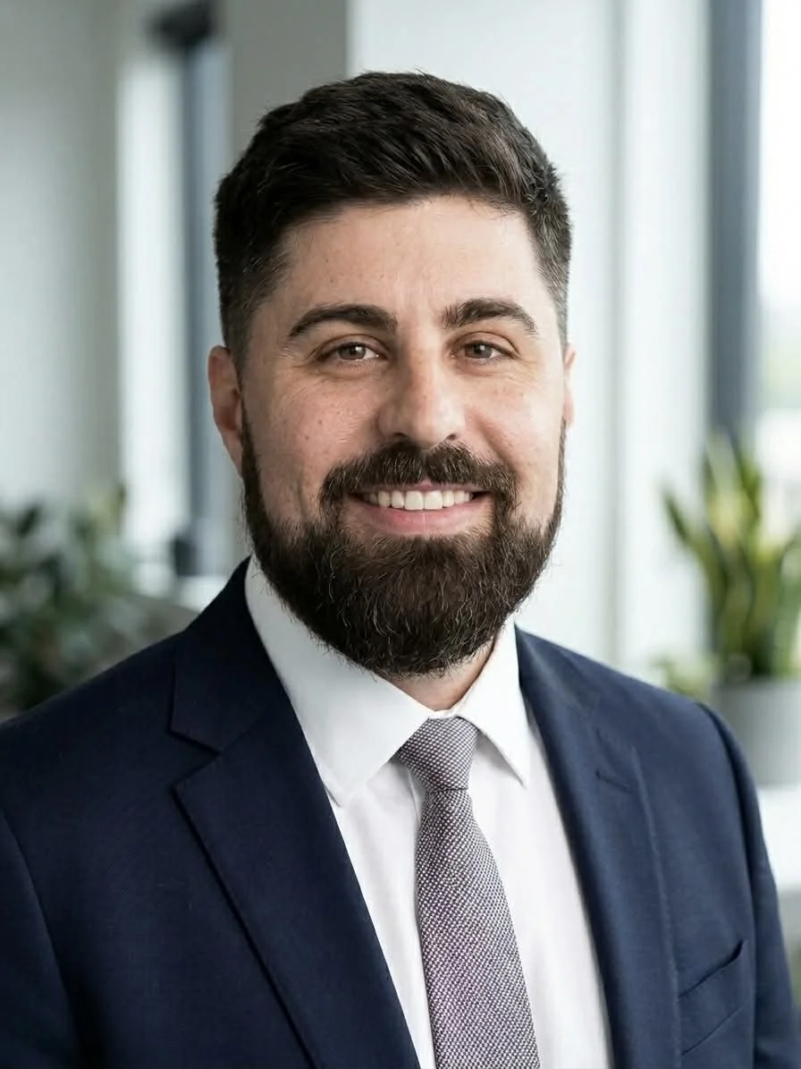A man with a beard and mustache wearing a navy suit, white shirt, and gray tie, smiling in an office setting with large windows and indoor plants in the background.