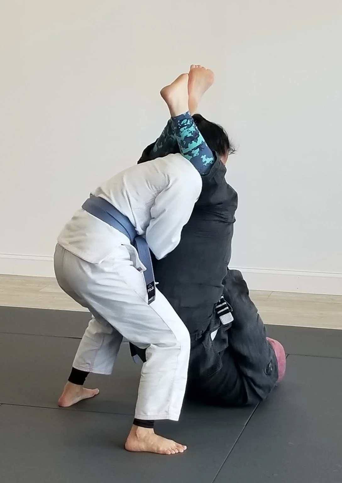 Two individuals practicing Brazilian Jiu-Jitsu on a black mat. One in a white gi with a gray belt is applying a submission hold on the other person wearing a black gi with a blue and black camo rash guard. The person in the white gi is in a crouching stance with one knee on the ground and one foot flat on the floor, focusing on maintaining the hold.