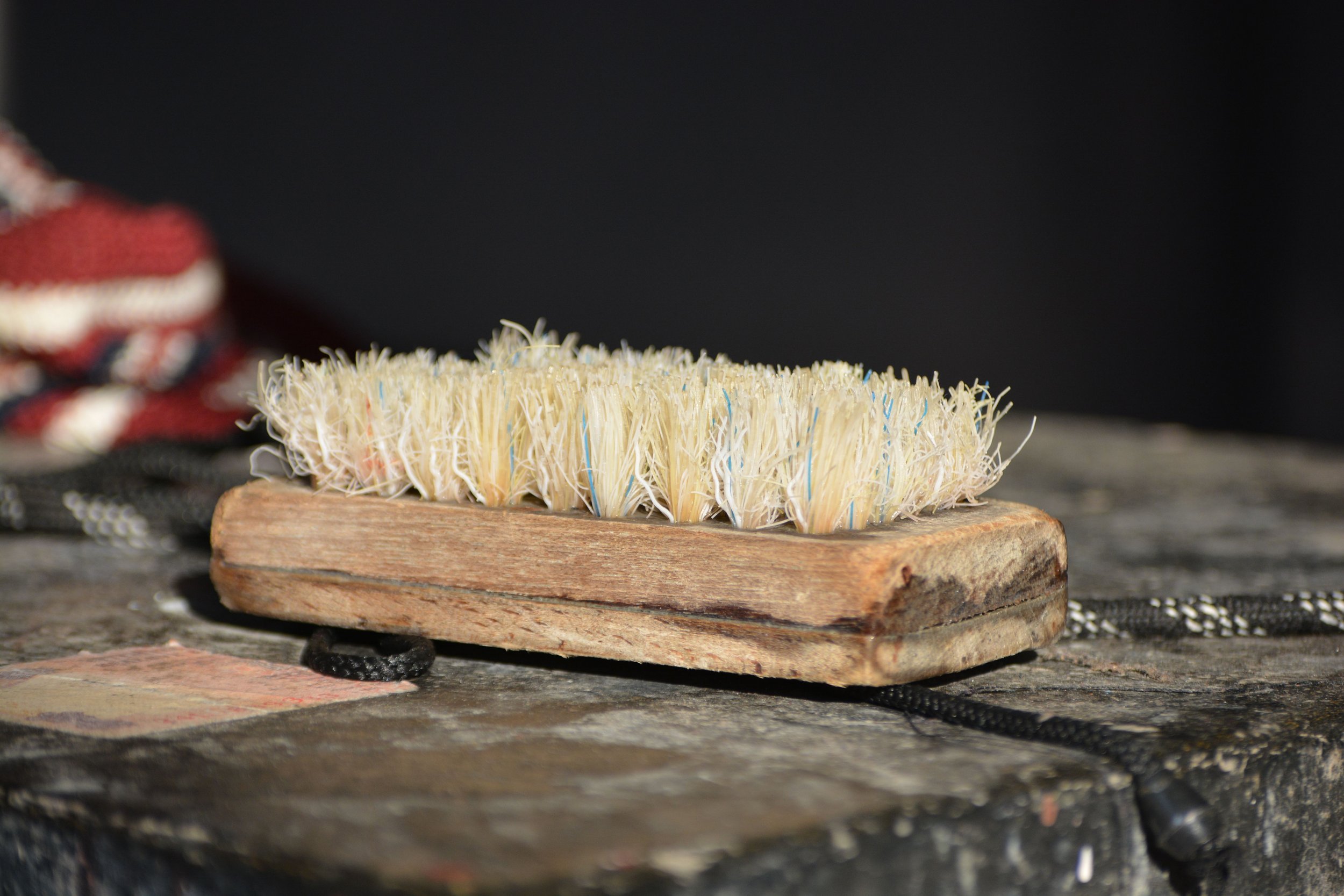 A cleaning scrub brush with white bristles and blue accents on a wooden base. The brush is placed on a rustic, weathered wooden surface.