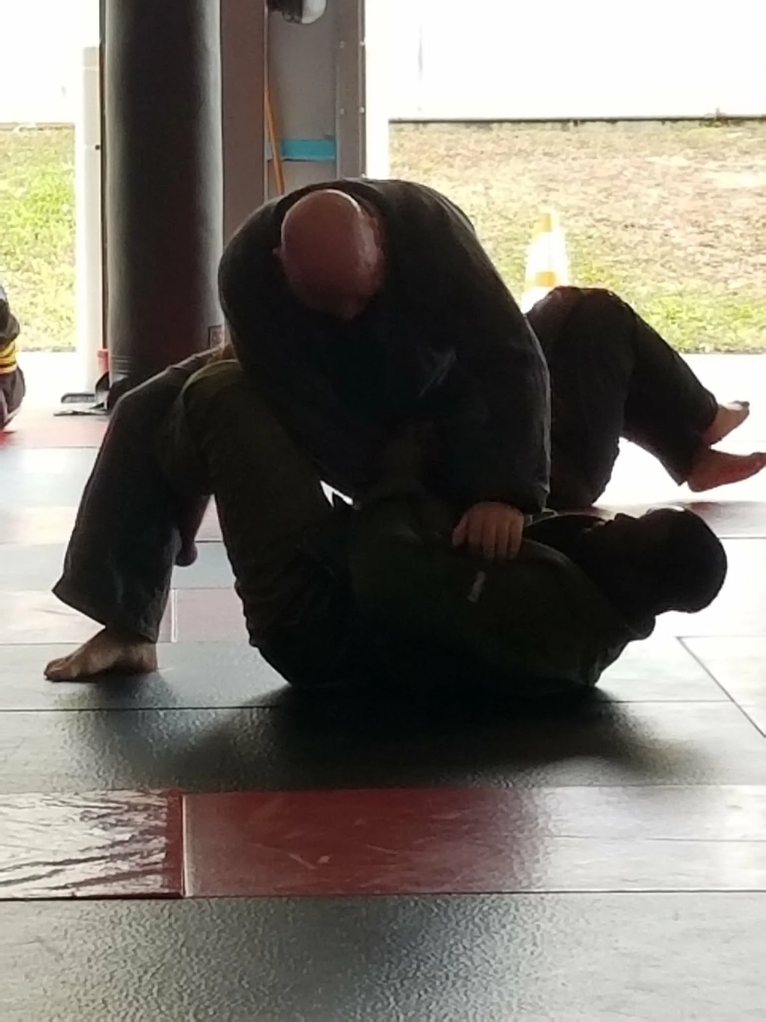 Two martial artists in black gi uniforms practicing Brazilian Jiu-Jitsu on a red and black mat in a gym with an open garage door.