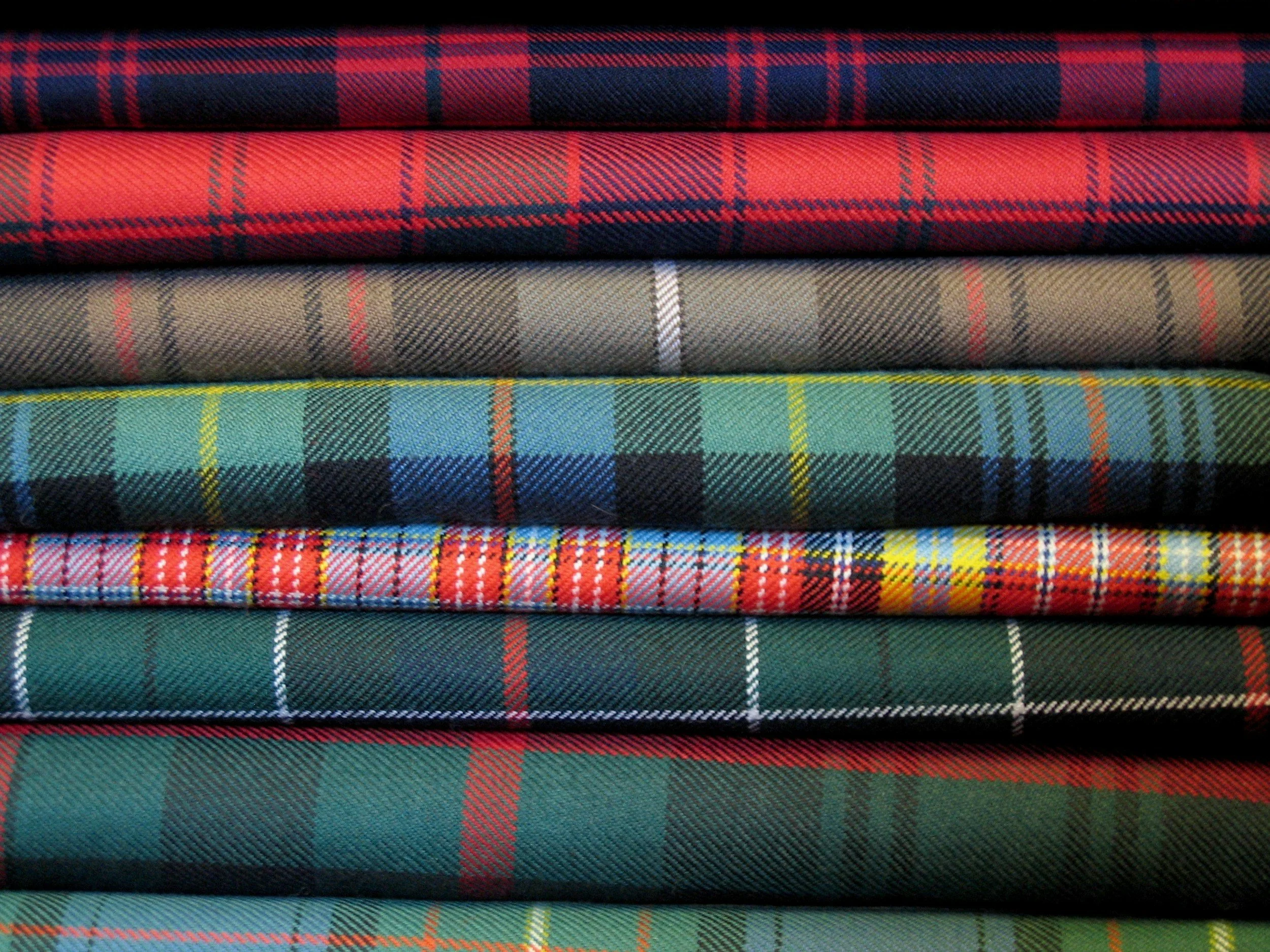 Stacked folded plaid fabric in various colors and patterns, including red, blue, green, beige, and yellow with different plaid designs.