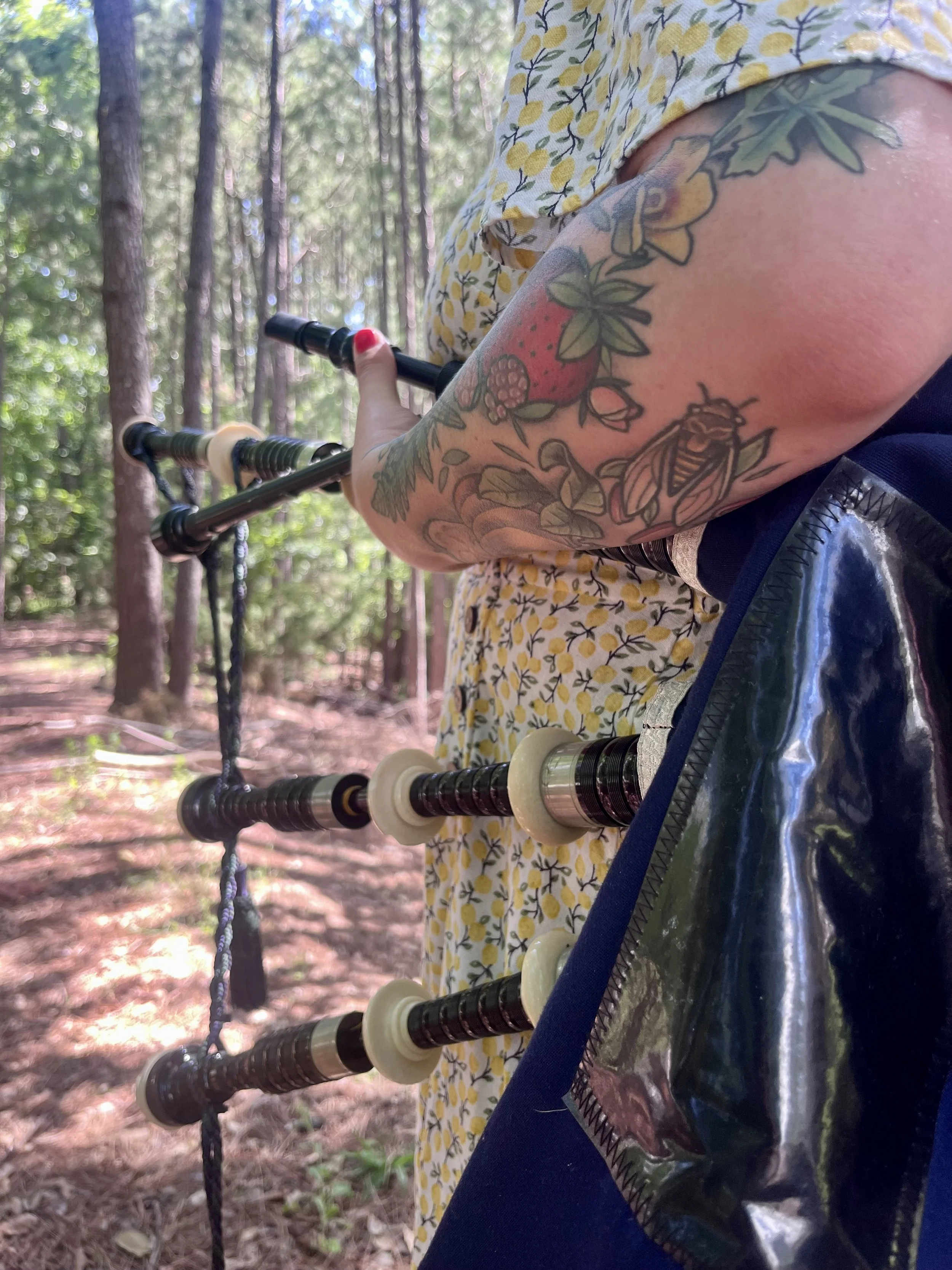 A person with floral tattooed arm holding a set of black and white bagpipes in a forest with trees and sunlight.