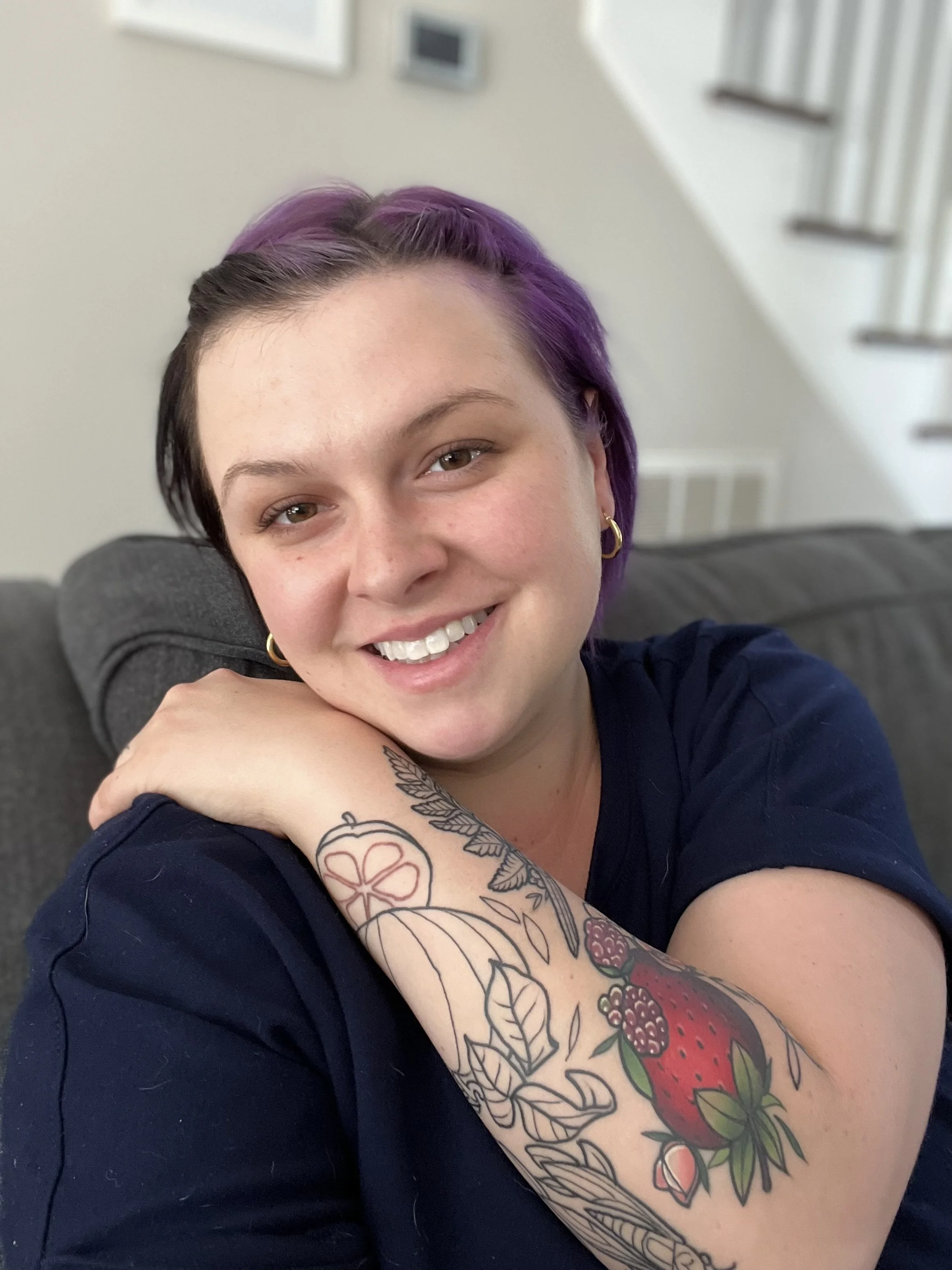A woman with short purple hair and piercings smiling, resting her head on her arm, which has a colorful tattoo of lemons, strawberries, and leaves, while sitting on a gray couch in a room with a staircase and white walls.