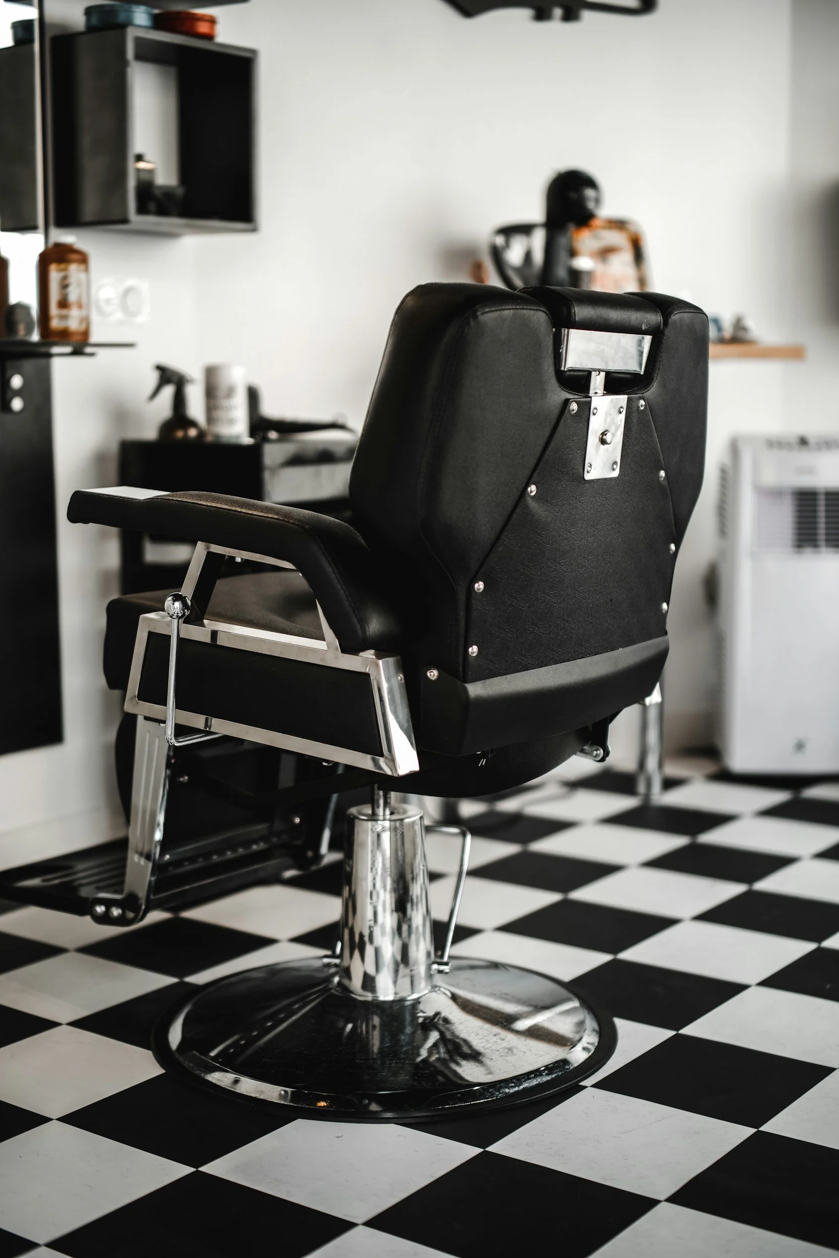Empty black barber chair in a barbershop with black and white checkered floor, shelves with grooming products, and a mirror in the background