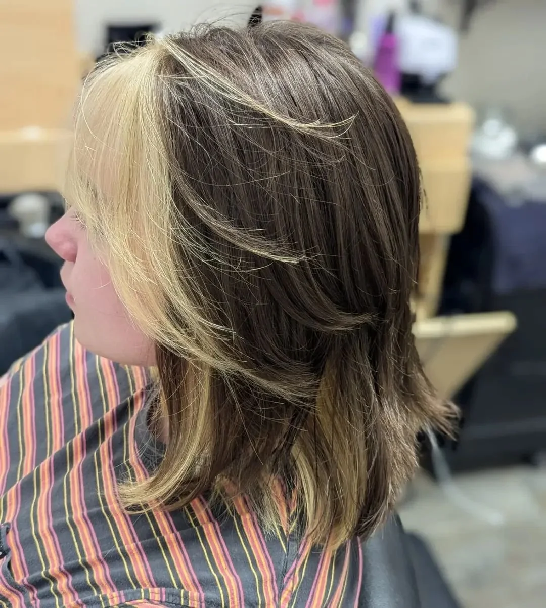 Side view of a woman with shoulder-length layered brunette hair with blonde highlights in a salon setting.