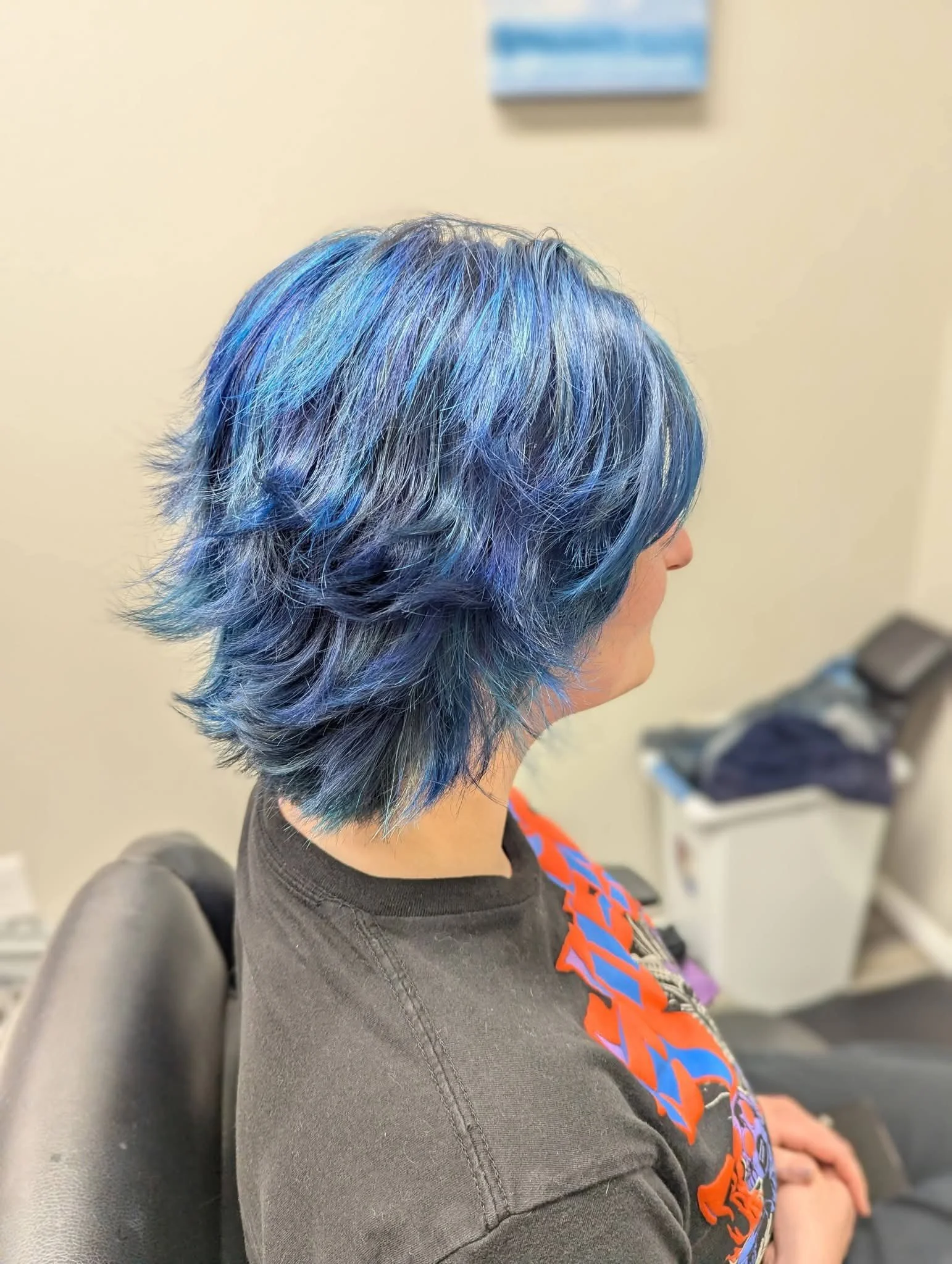 Profile of a person with short, layered hair dyed in shades of blue, purple, and gray, sitting in a waiting room with beige walls and a blurred blue sign in the background.