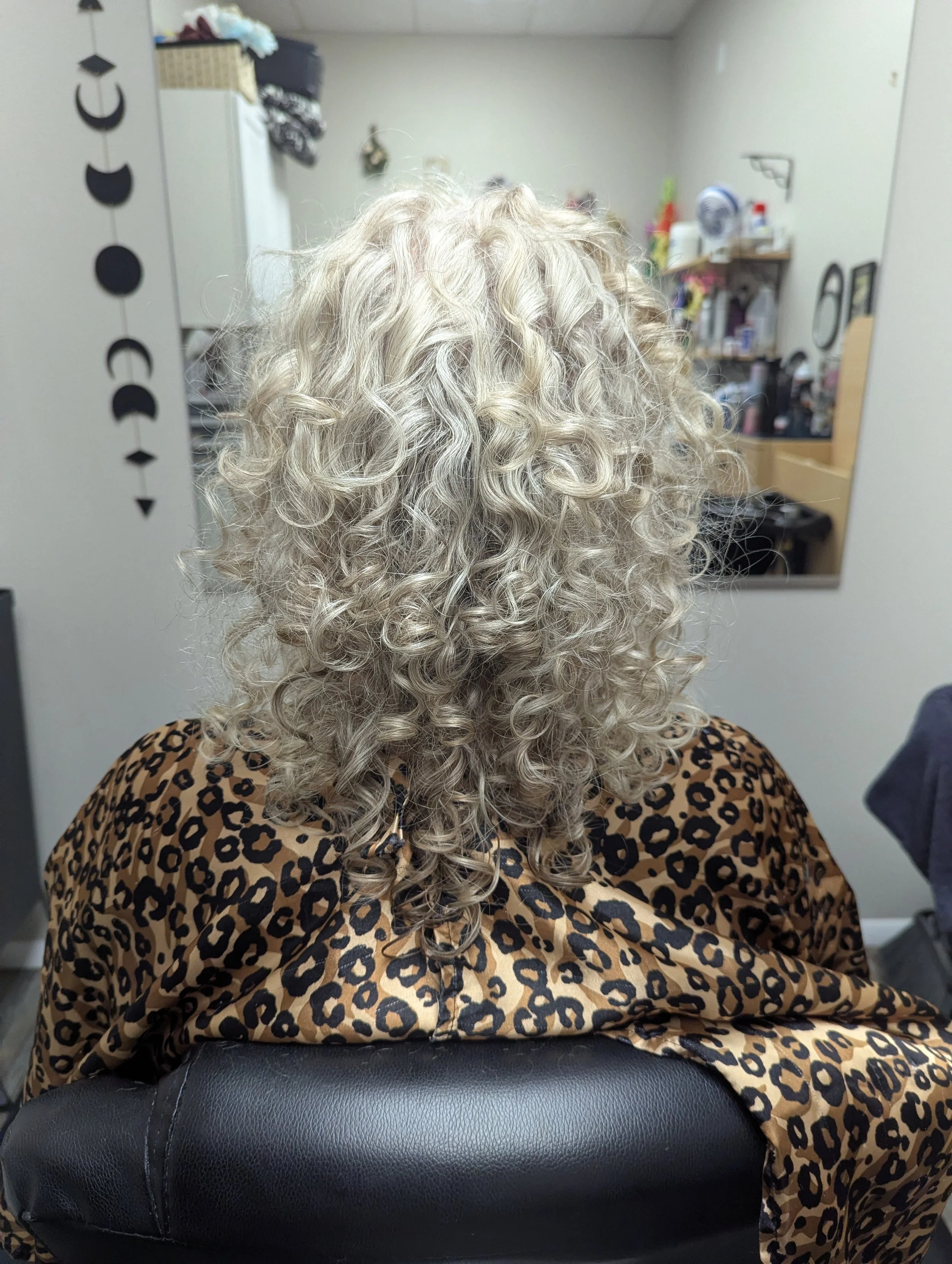 Back of a person with long, curly, platinum blonde hair wearing a leopard print top, sitting in a salon or hairdresser's chair, with a wall-mounted mirror and salon supplies in the background.