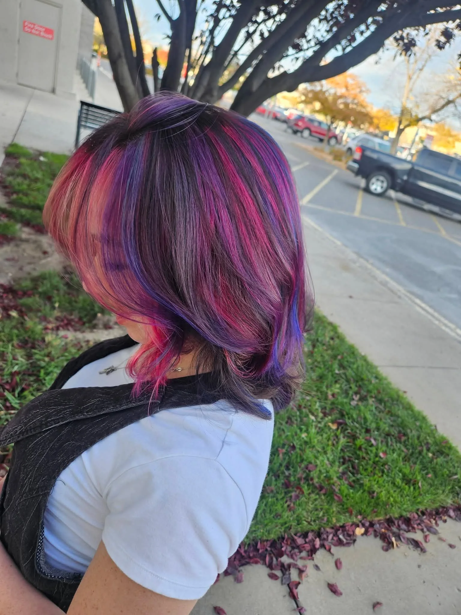 A person with vibrant, multi-colored hair in shades of purple, pink, orange, and blue stands outdoors near a tree with autumn leaves, wearing a white shirt and a black backpack.
