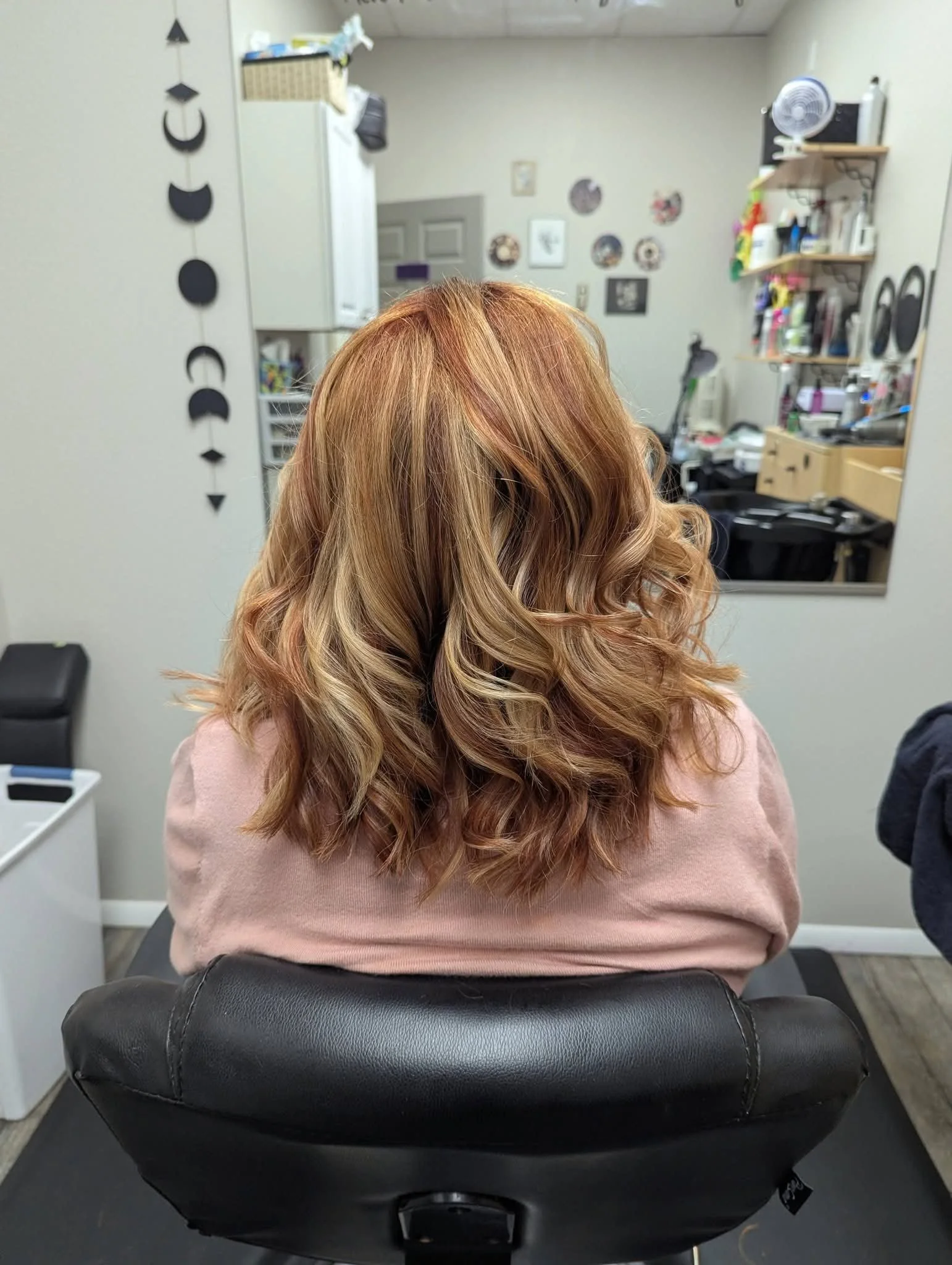 Back view of a woman with long, wavy, reddish-blonde hair sitting in a salon chair, framed by a large mirror with a wall decorated with small picture frames and a shelf holding hair products and tools.
