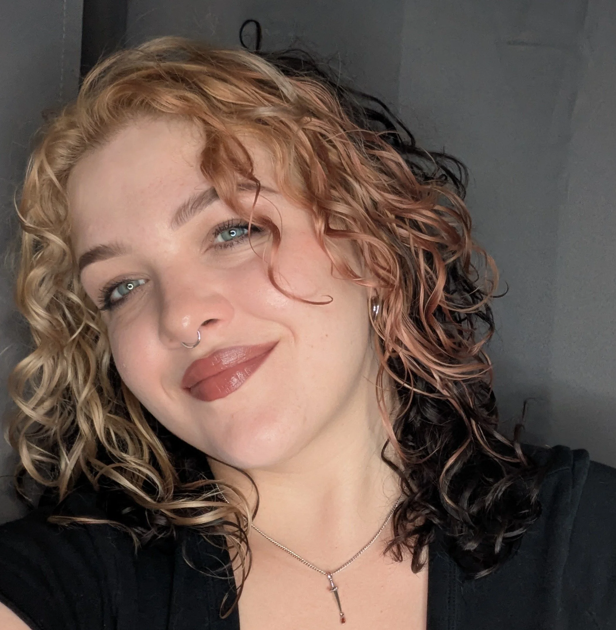 Close-up of a woman's face with curly hair, blue eyes, wearing a necklace with a pendant and earrings, against a dark background.