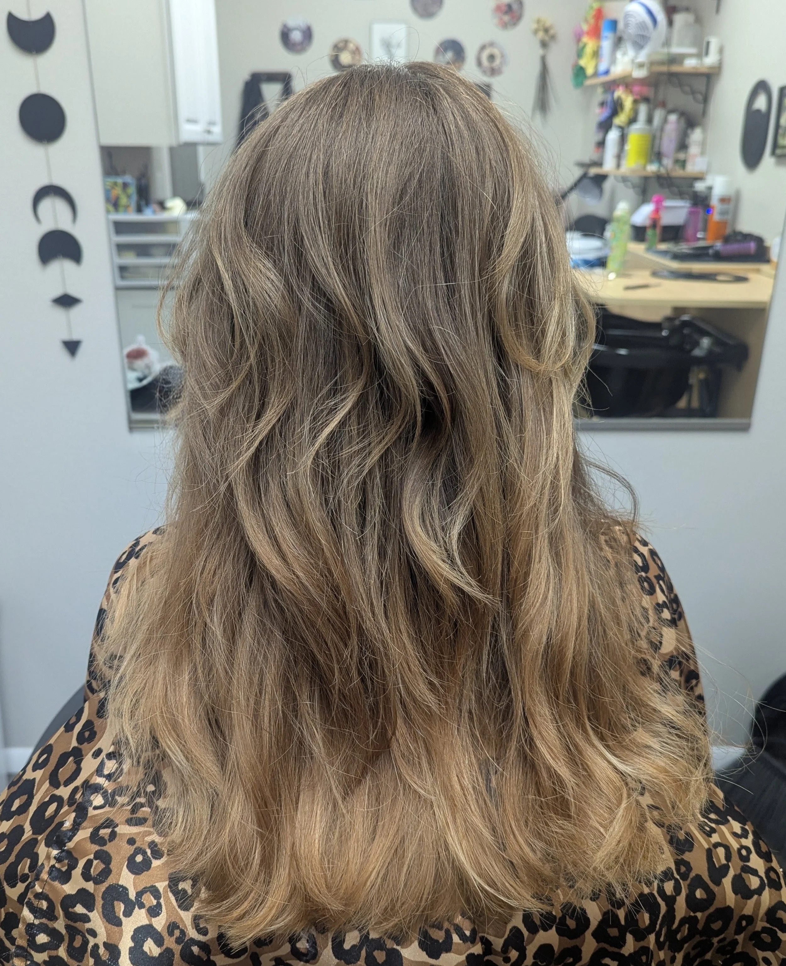 Close-up of a person's head with long, wavy, light blonde hair in a salon, with styling products and tools on shelves behind.