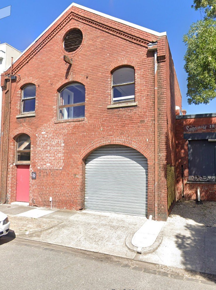 A red brick building with a large garage door, a pink front door, and three arched windows, under a clear blue sky, with a white car partially visible on the street.