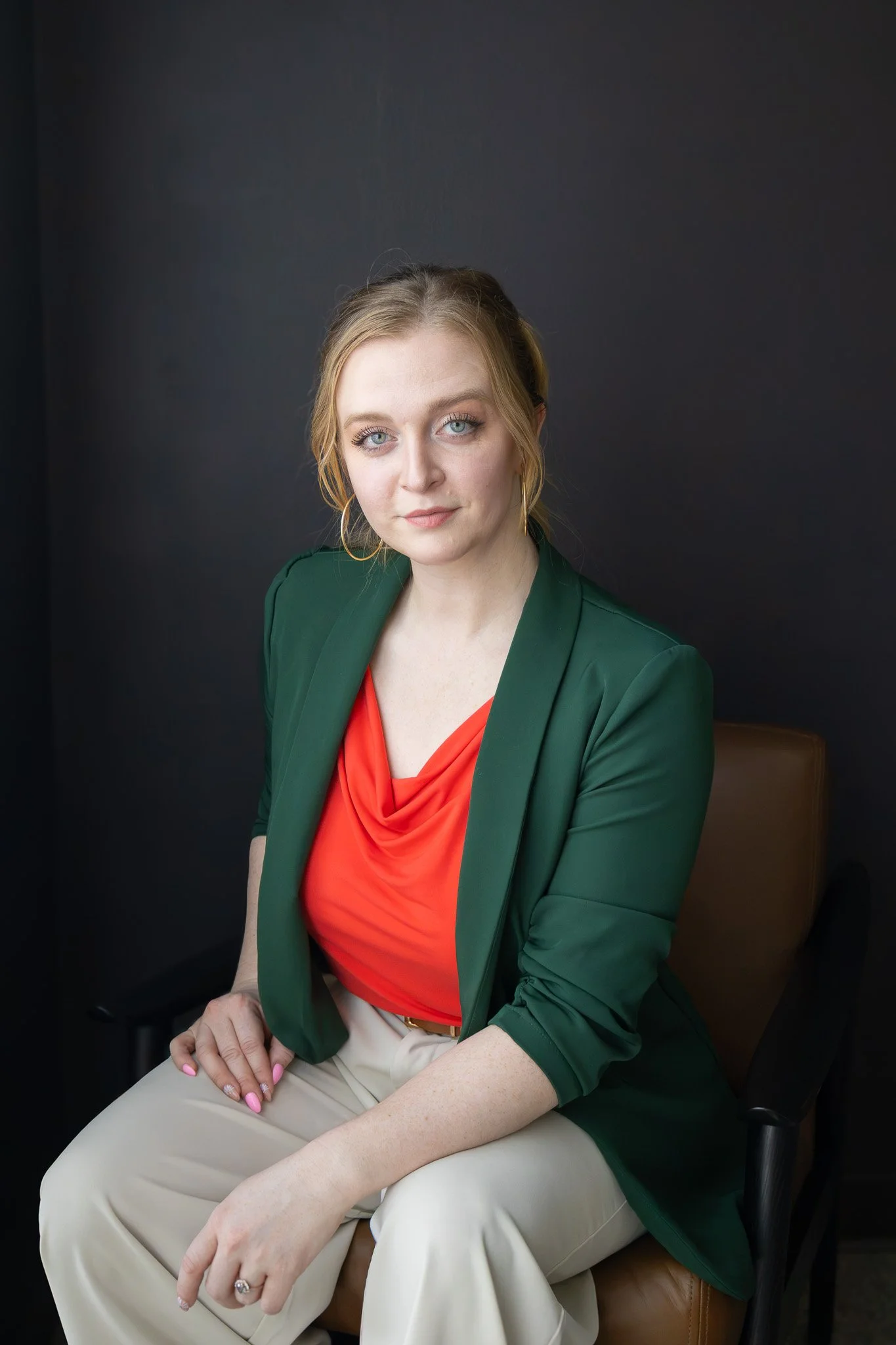 A woman sitting in a chair against a dark background, wearing a green blazer, red top, and beige pants, with blonde hair, green eyes, and hoop earrings.