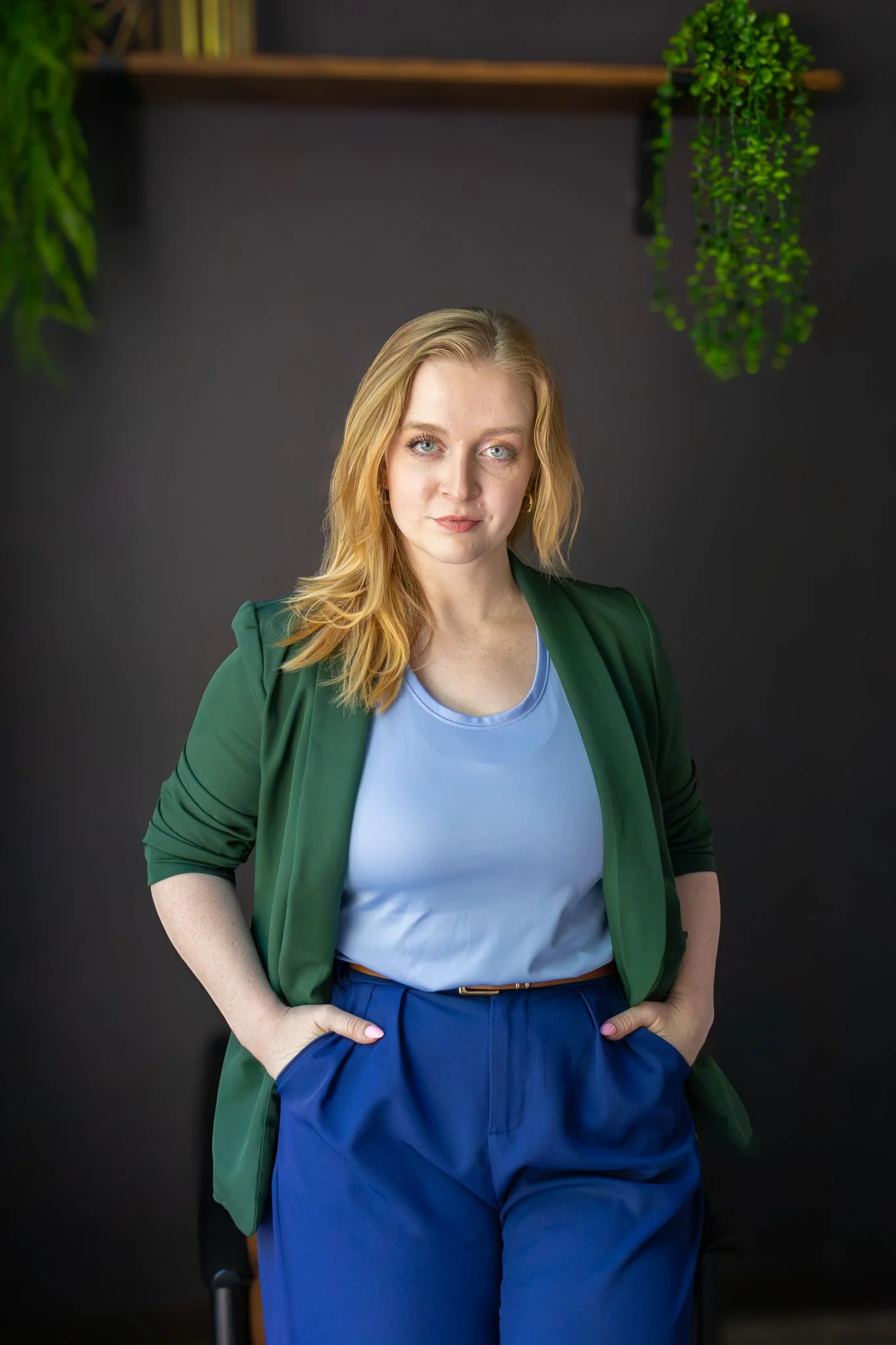 A woman with blonde hair, wearing a green blazer, light blue shirt, and blue pants, standing with hands on hips in front of a dark wall with hanging green plants.