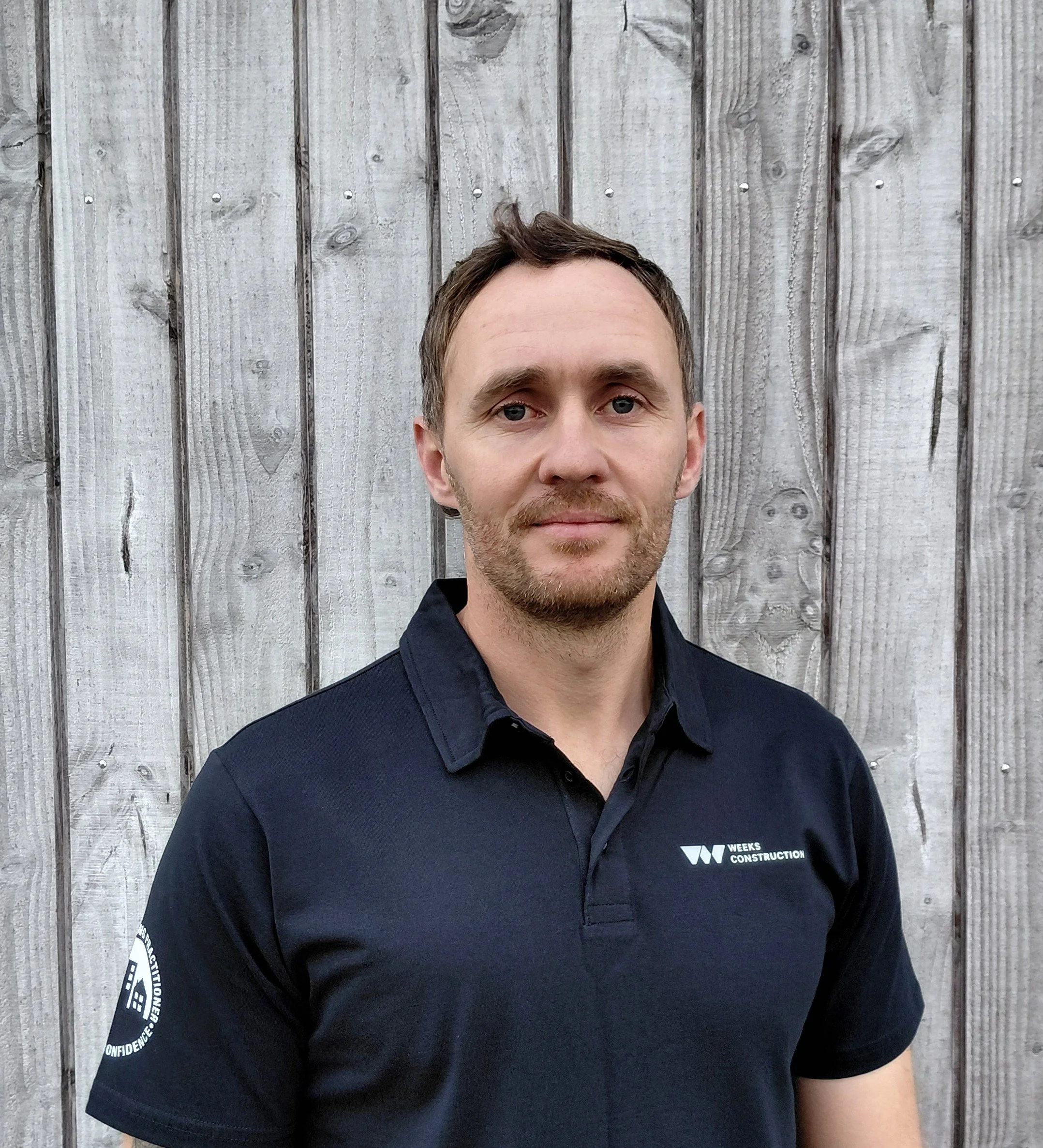 Close-up of a man wearing a black polo shirt with a logo on the chest, standing in front of a weathered wooden fence.
