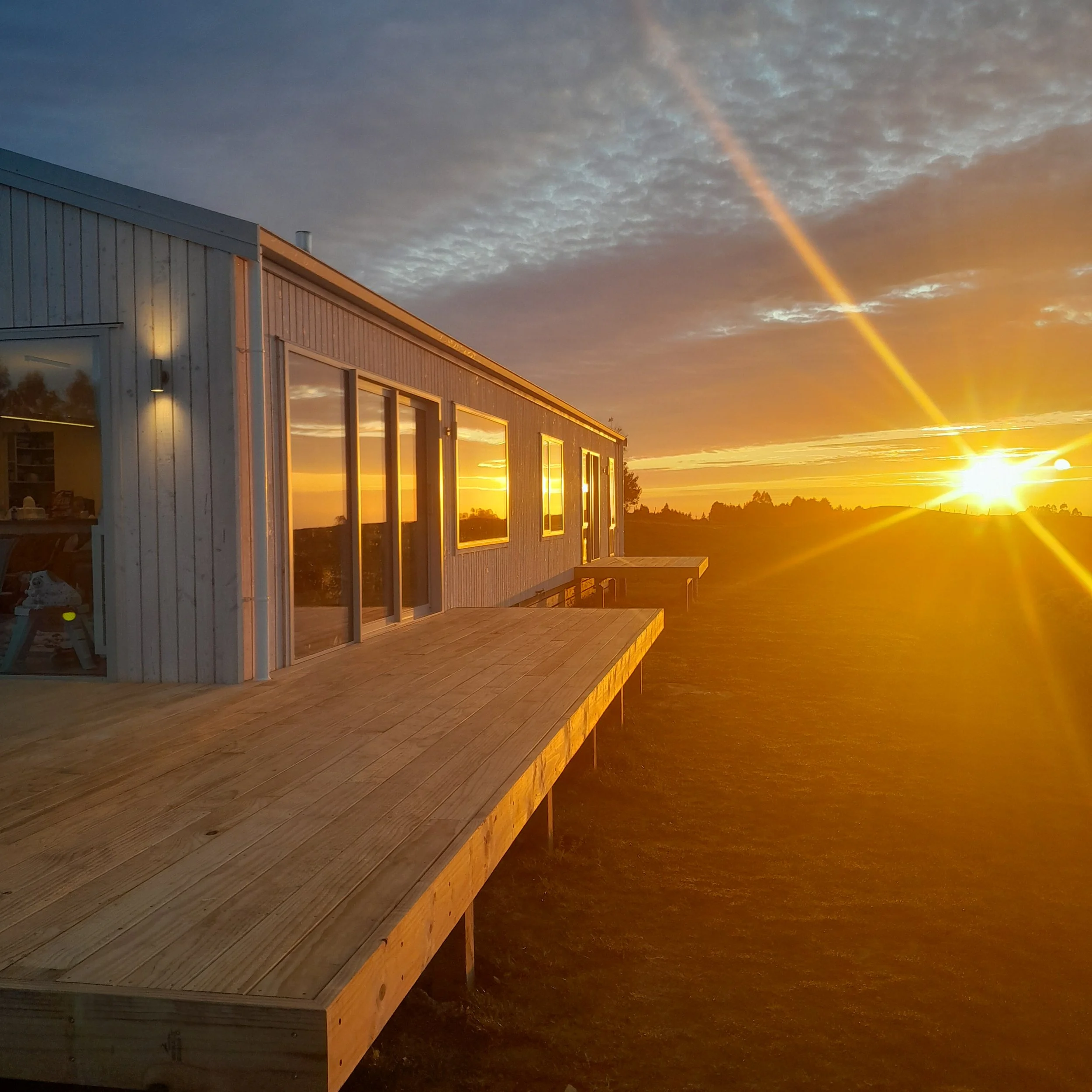 Sunset over a modern house with large windows, reflecting the orange and pink hues of the sky, with a wooden deck in the foreground and a vast open landscape.