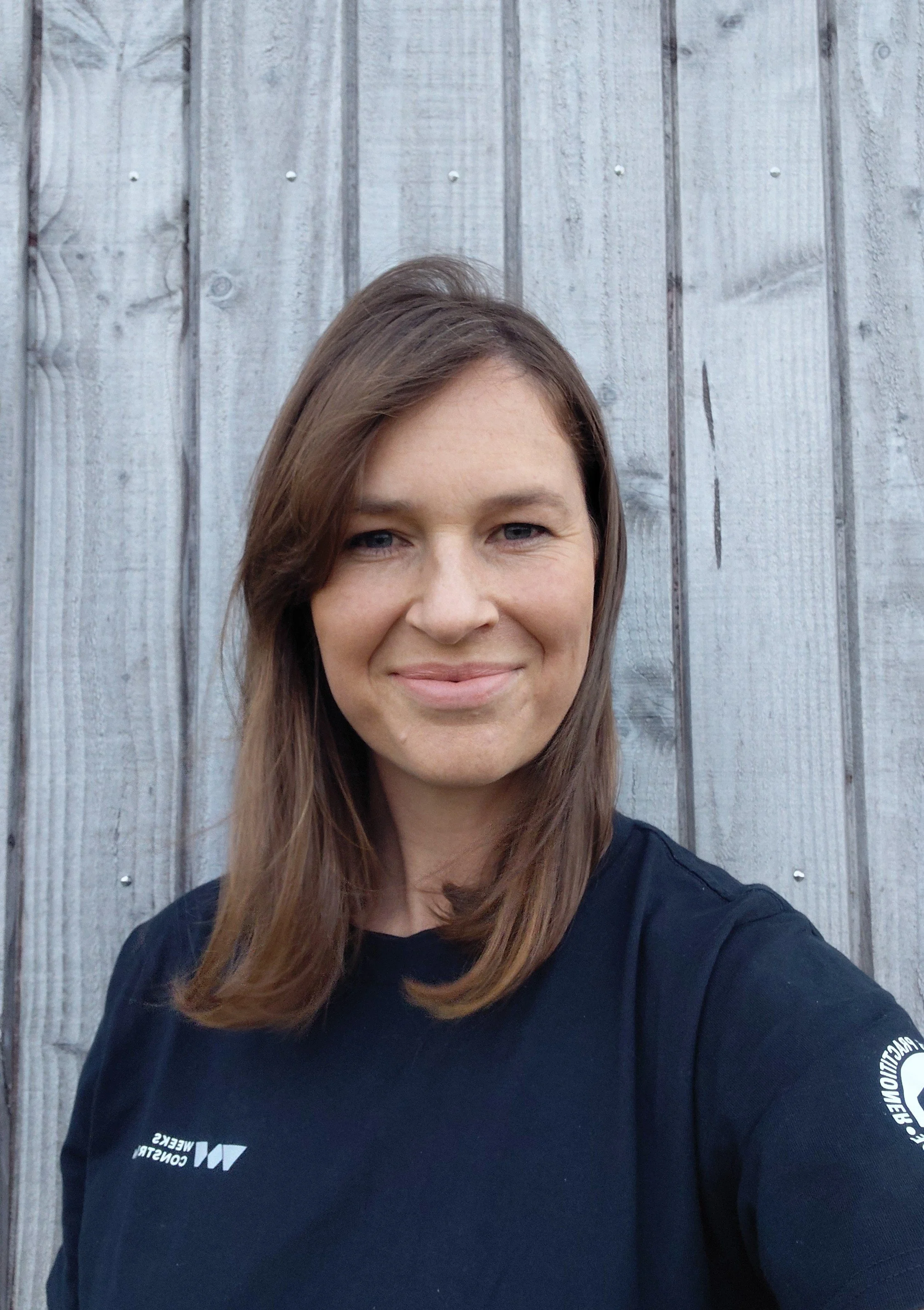 A woman with long brown hair wearing a black shirt standing in front of a weathered wooden fence, smiling at the camera.
